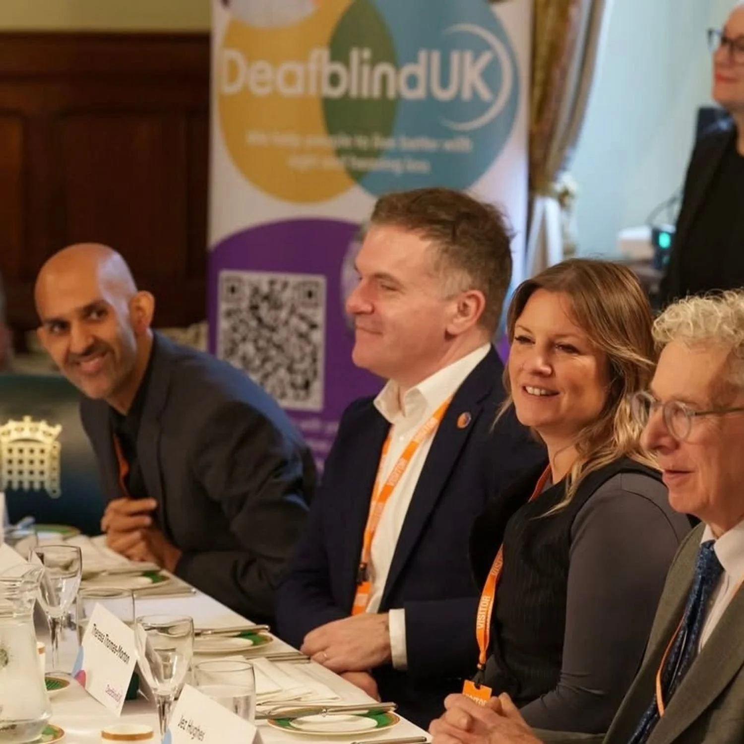Four people sitting at a conference table with glasses, plates, and name tags, smiling and engaging in a discussion. In the background, there is a banner with the logo and text for Deafblind UK.