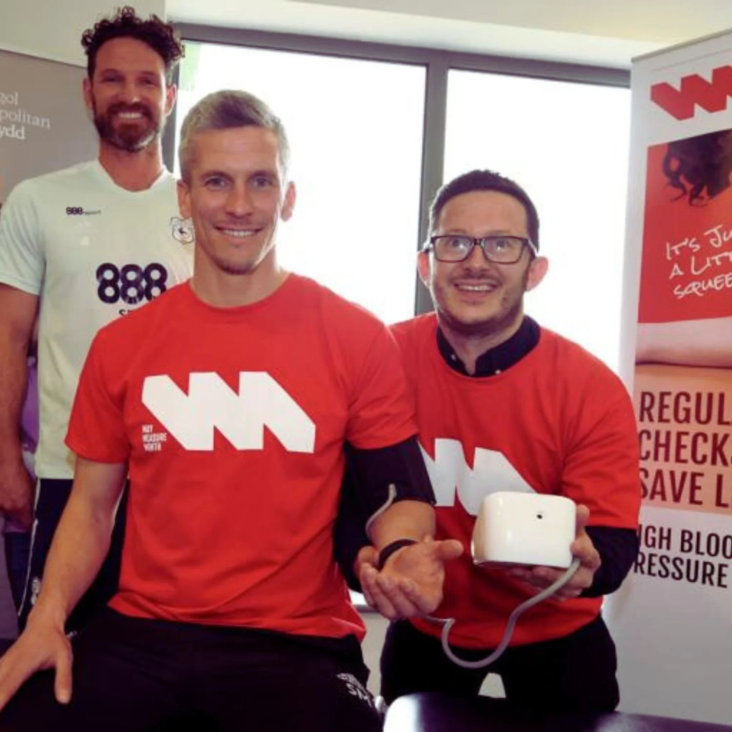 Three men smiling at a health event, two wearing red shirts with a white logo, one in the background wearing a white shirt with purple and black graphics. The man on the right is holding a blood pressure monitor.