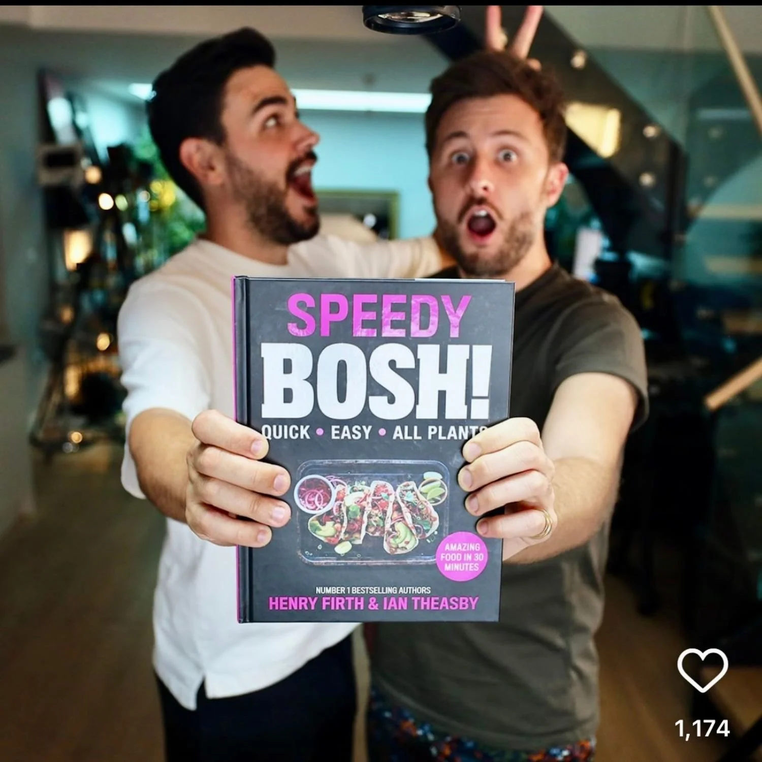 Two men are celebrating happily indoors, holding a cookbook titled 'Speedy Bosh!' with a subtitle 'Quick · Easy · All Plants.' One man is giving bunny ears to the other behind his head. The background shows a room with a staircase and some plants.