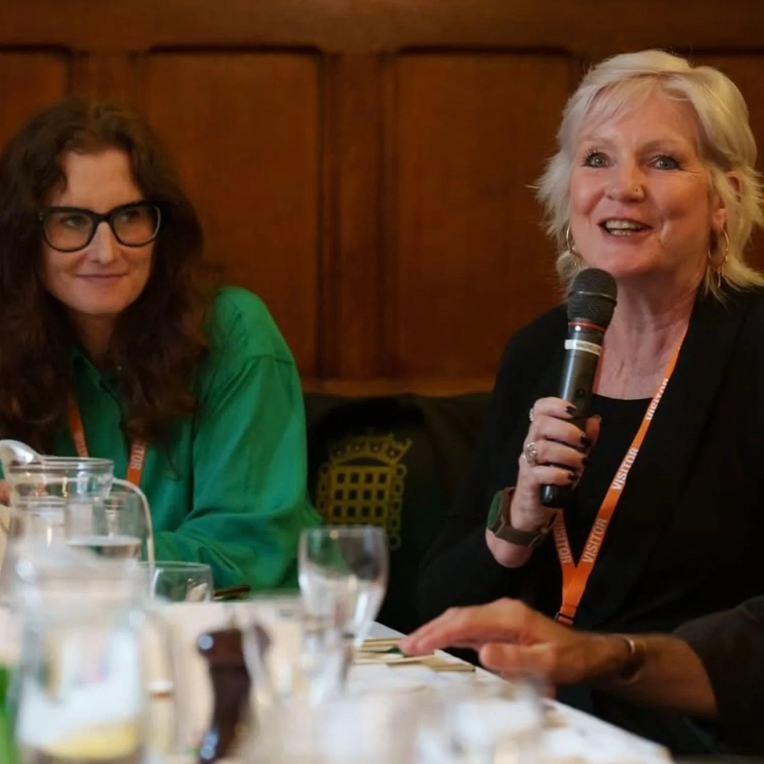 Two women sitting at a table during a panel discussion or meeting, one speaking into a microphone, with glasses of water and other beverages on the table.