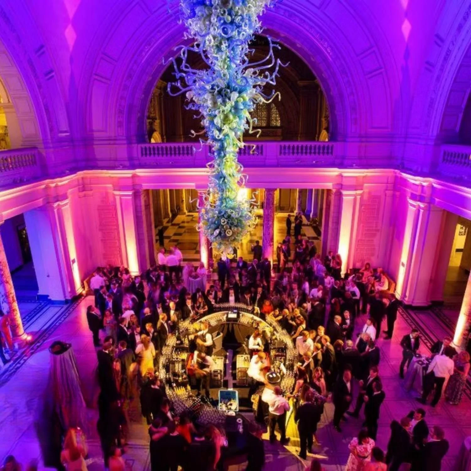 A large, elegant indoor event with colorful lighting, a central floral chandelier, and a crowded circular bar surrounded by guests in formal attire.