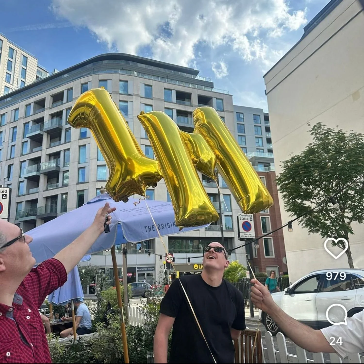 Three people celebrating outdoors with large gold balloons.  One person is holding the balloons, another is smiling and looking up, and the third is holding a string. There are buildings, trees, and a busy street in the background.