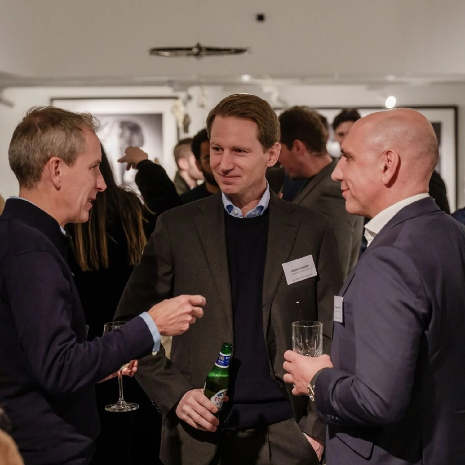 Three men are engaged in conversation at a networking event with drinks in hand. The man on the left is speaking and holding a wine glass, the man in the middle has a beverage bottle and is listening, and the man on the right holds a glass and listens.