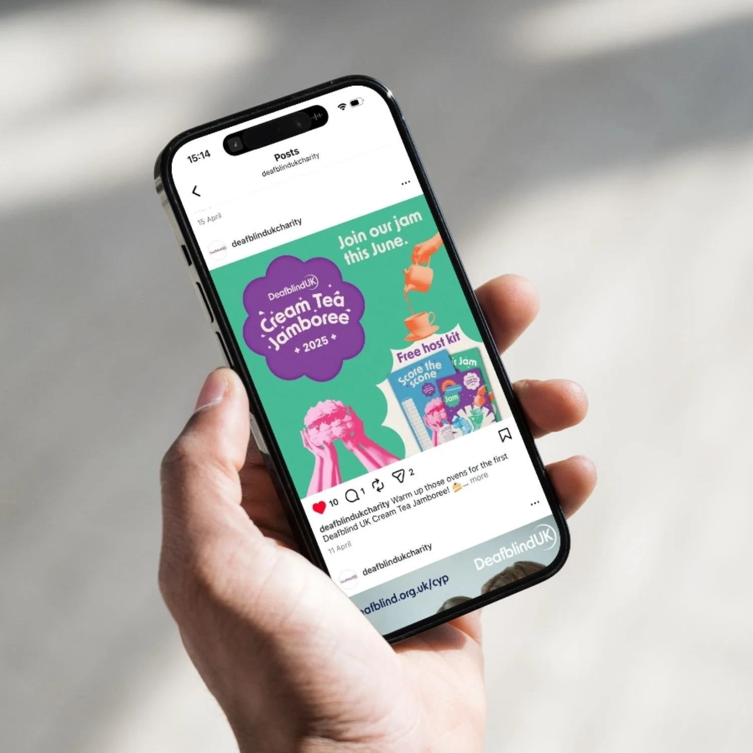 A person holding a smartphone displaying a colorful social media post about a cream tea jamboree event in June, with images of hands holding a cake, a teacup, and event details.