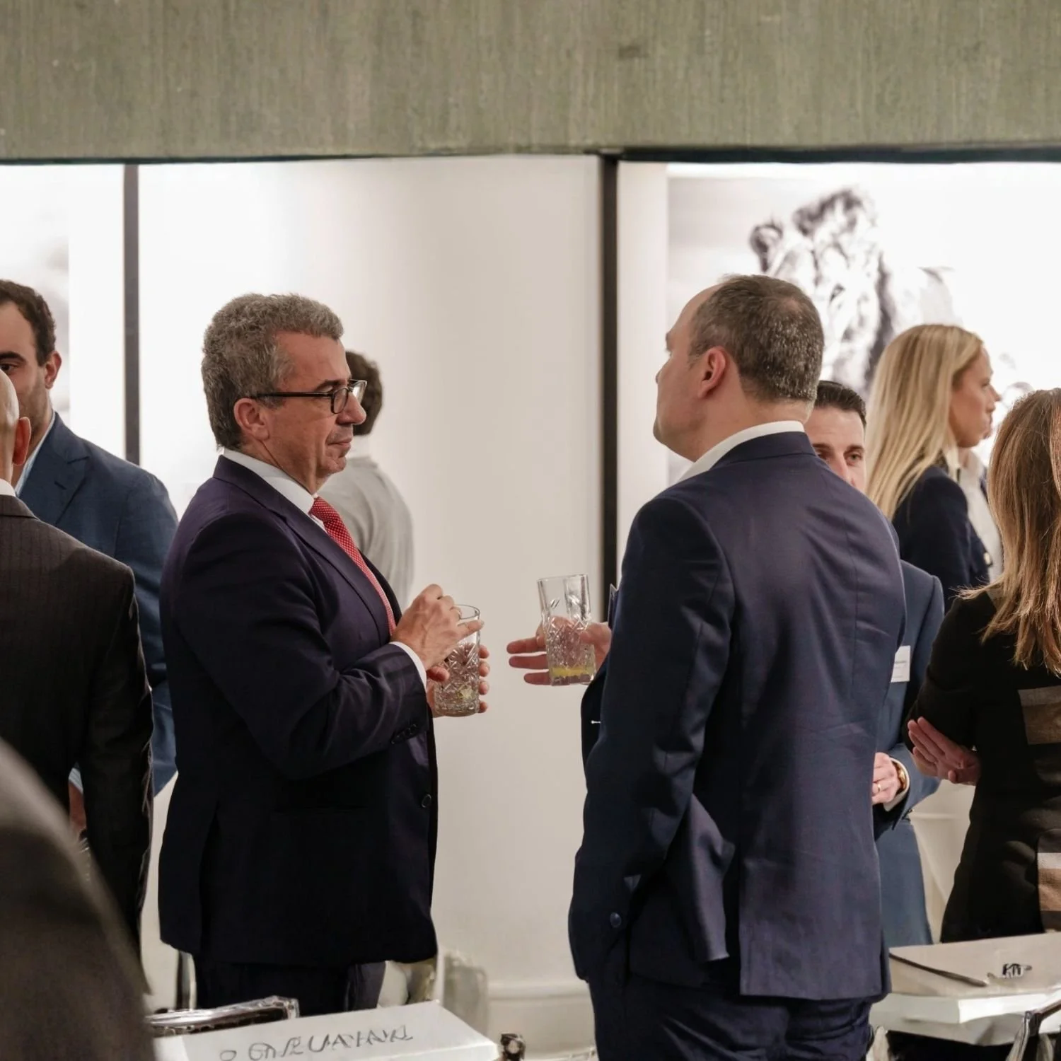 Two men in suits talking and holding glasses of drinks at a networking event, with other people socializing in the background.