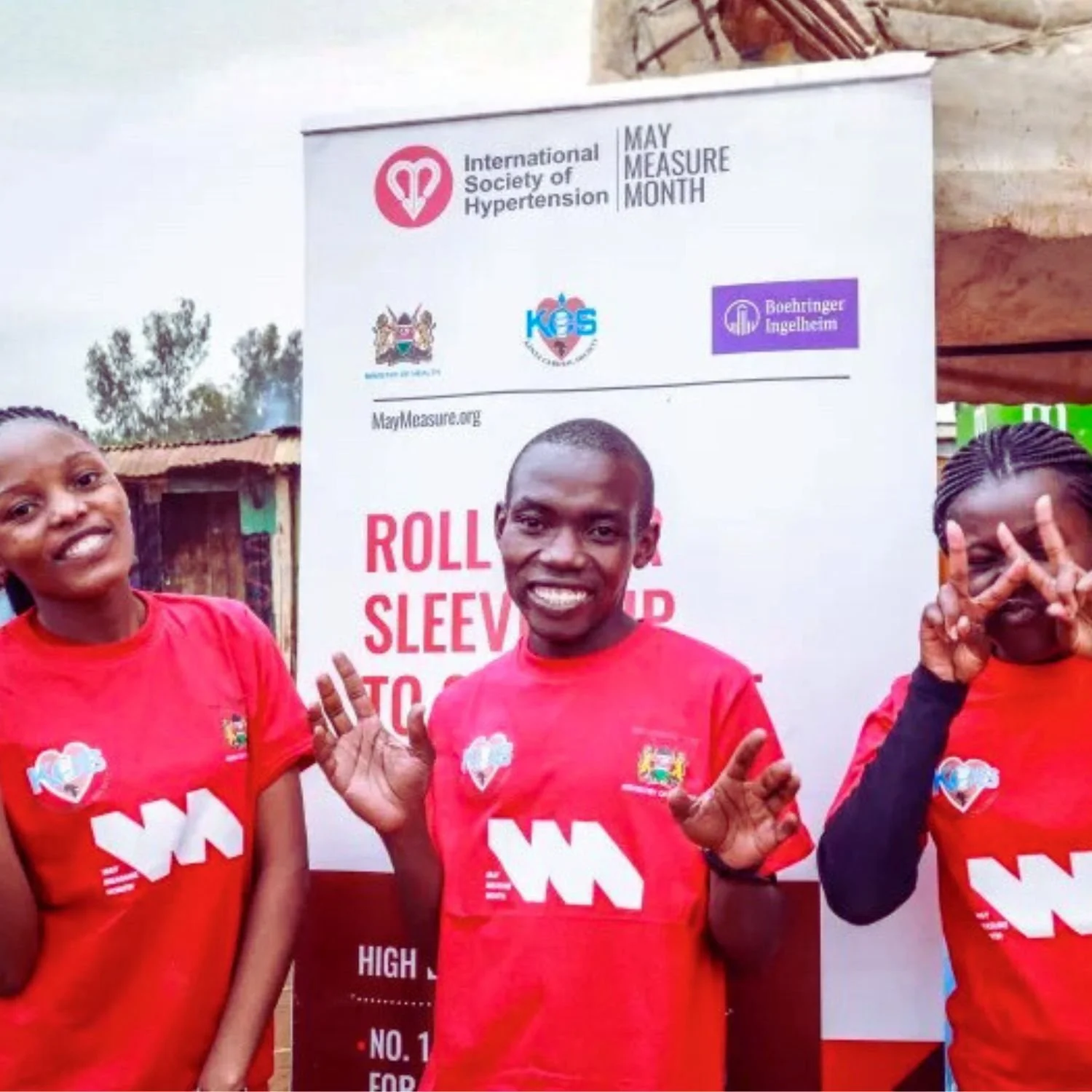 Three people wearing red shirts with logos, smiling and posing at a health awareness event for hypertension during May Measurement Month, with a banner in the background displaying health organization logos.