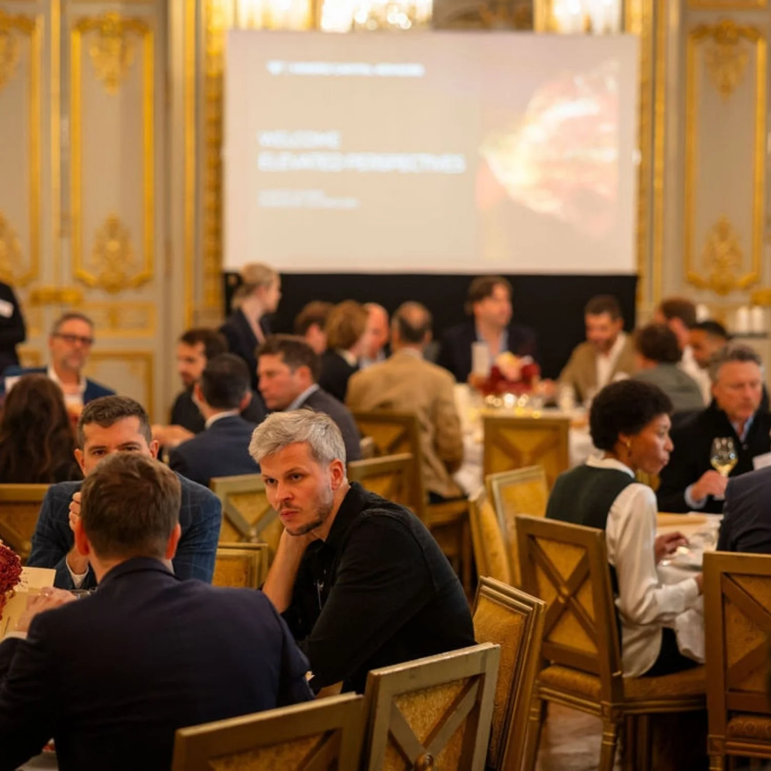 People sitting at tables in a grand, ornate room attending a conference or event, with a large screen on the wall displaying a presentation.