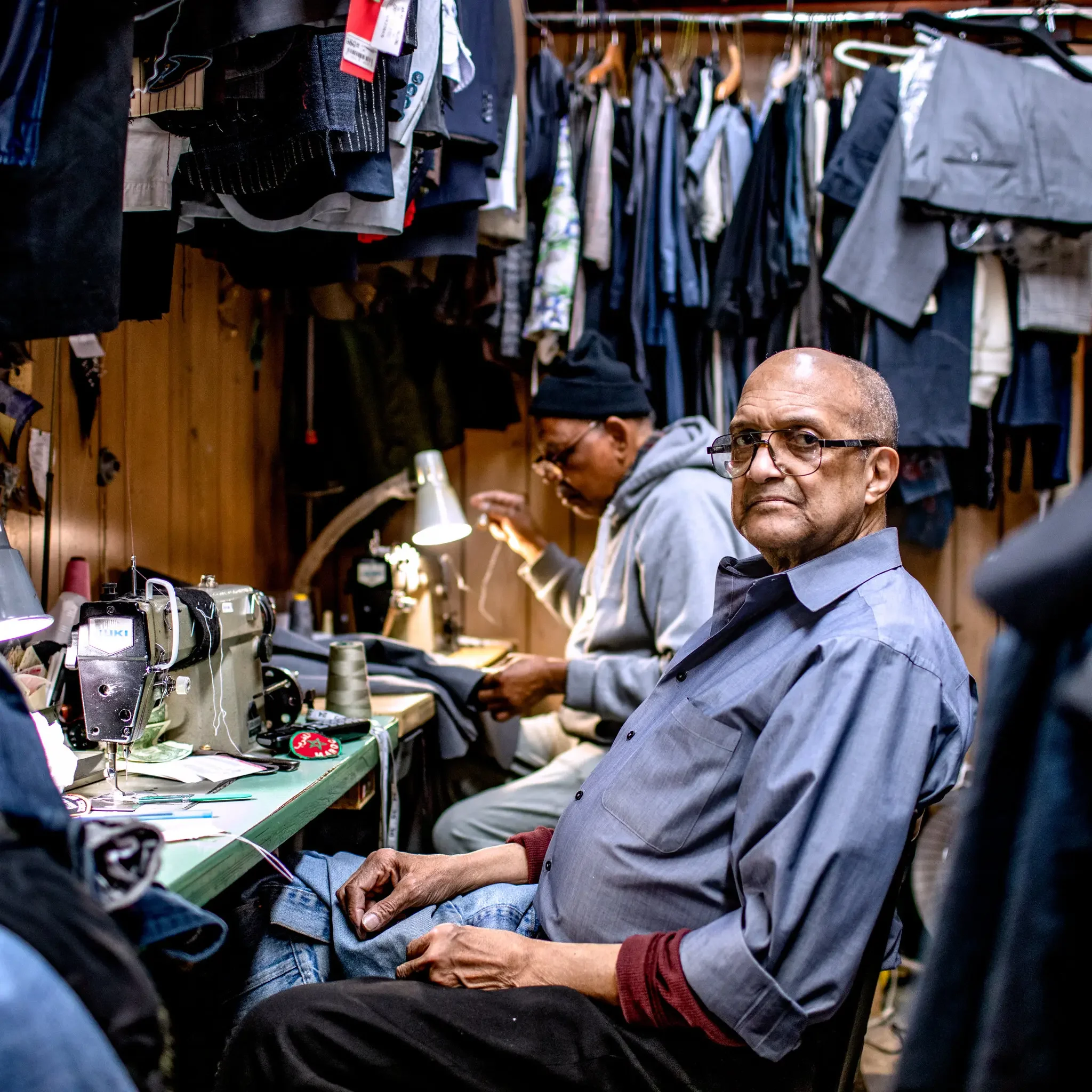 Two men working at a sewing station inside a clothing repair shop, surrounded by hanging clothes and fabric.