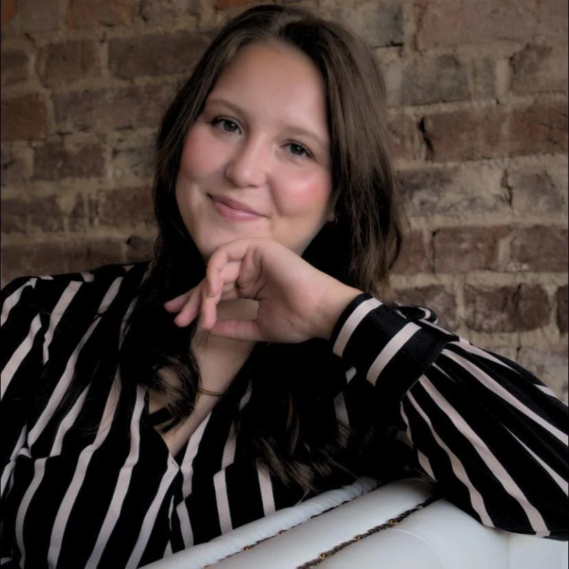 A young woman with long brown hair and a striped black and white shirt, smiling softly, sitting in front of a brick wall.