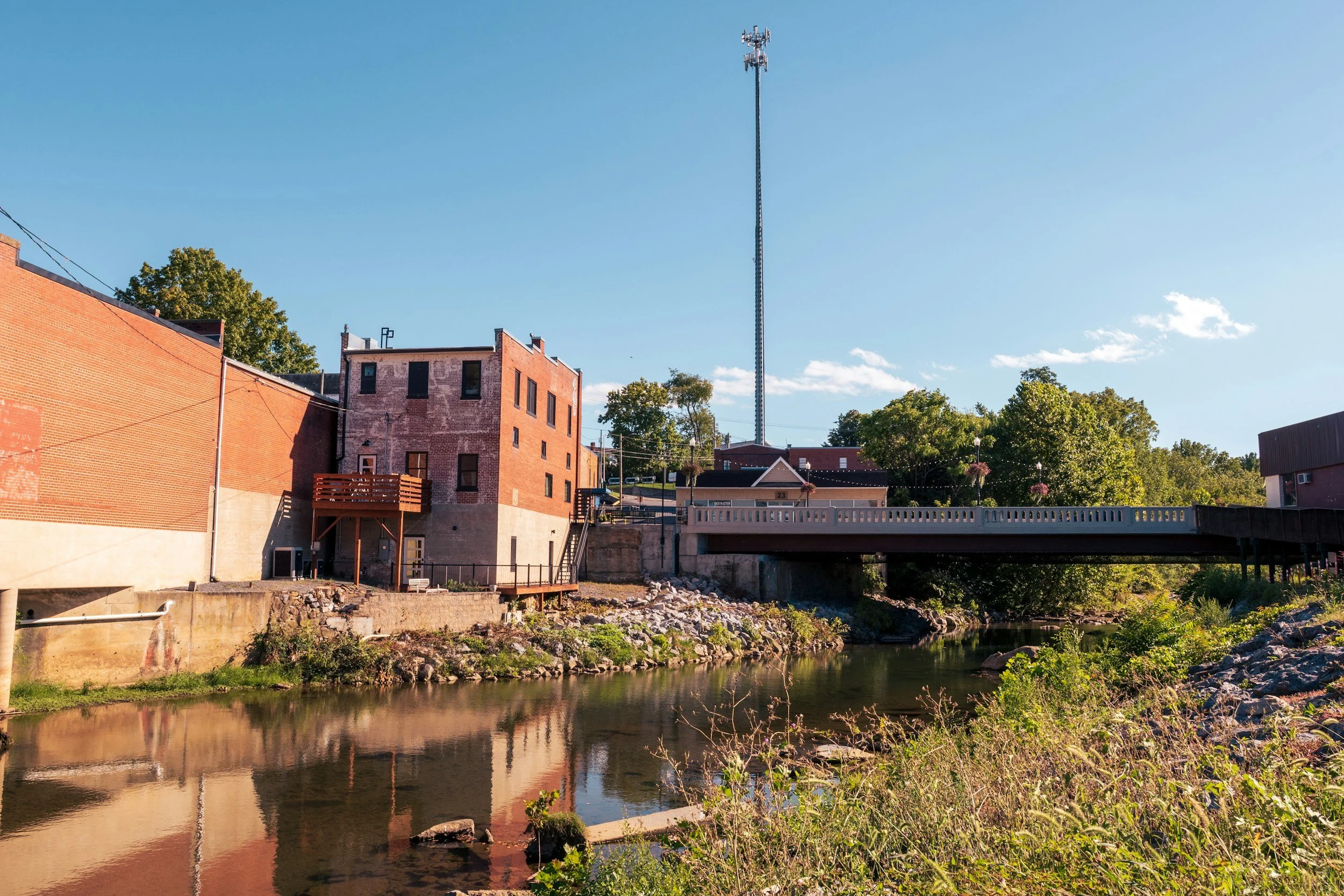 View of a river with rocks and greenery along the banks, under a bridge connecting brick buildings, with an old brick building and a tall communication tower in the background, and a clear blue sky.