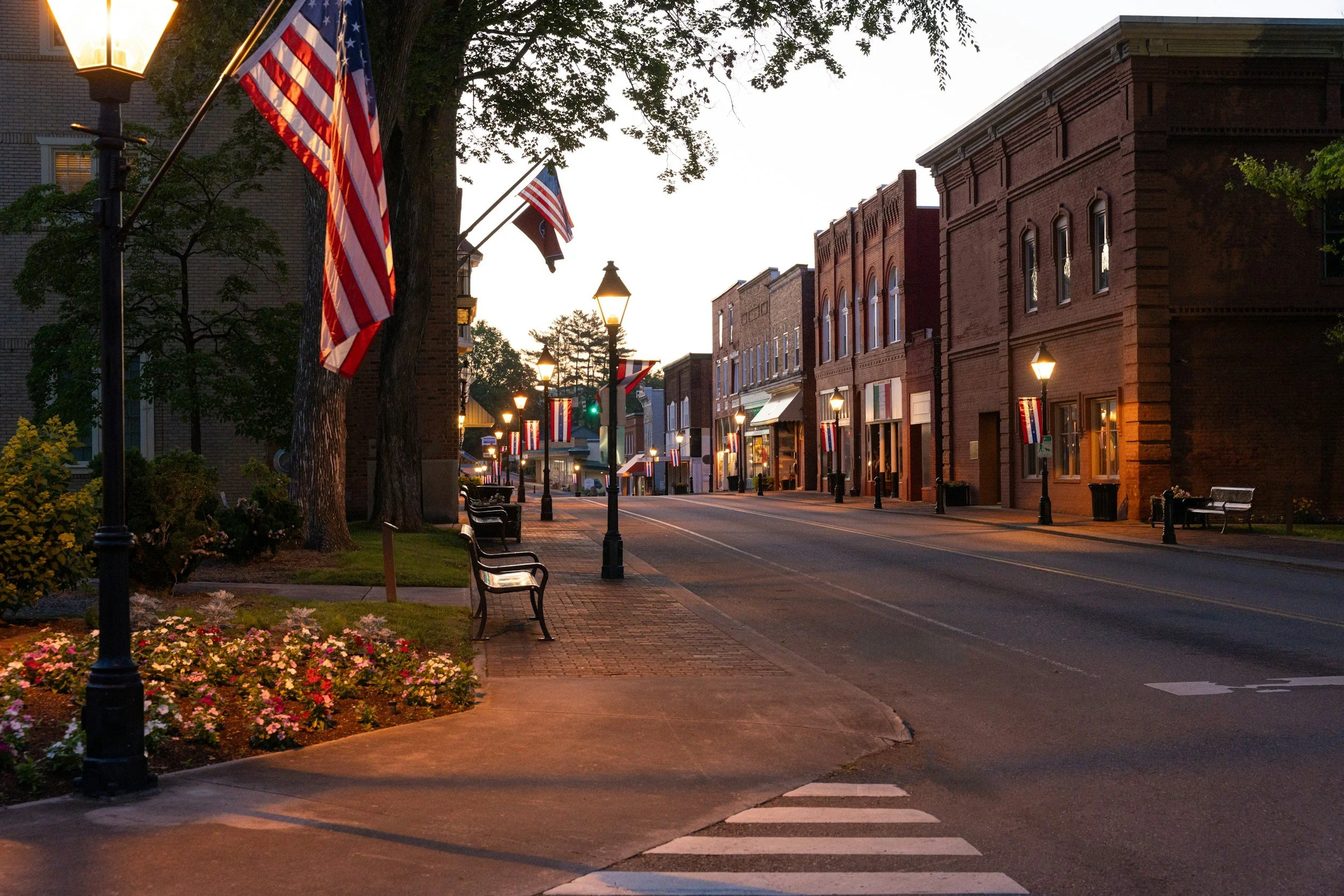 A quiet city street at dusk decorated with American flags, lined with brick buildings, street lamps, benches, and flowers.