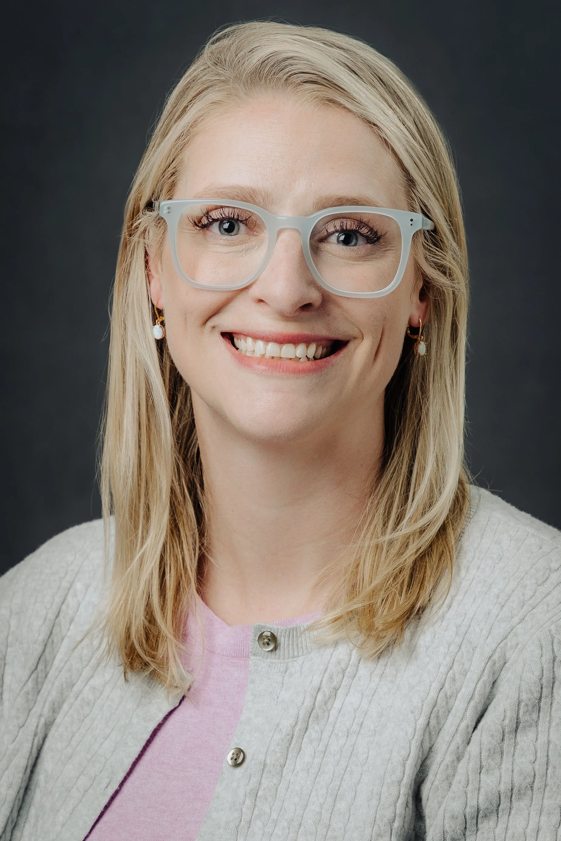 Smiling woman with blonde hair, wearing glasses, earrings, a pink top, and a light gray cardigan against a dark background.