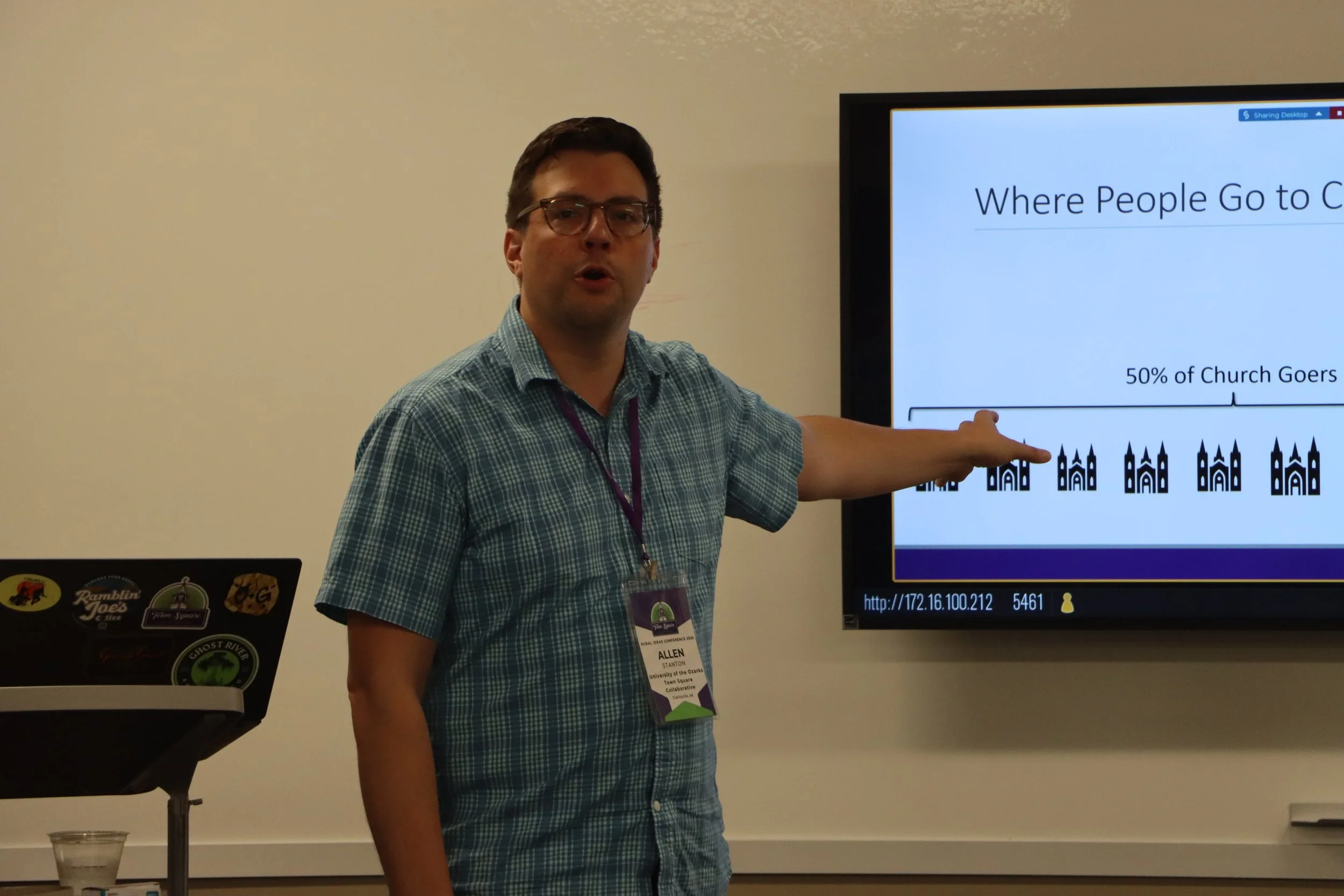 A man wearing glasses and a blue checkered shirt is giving a presentation, pointing towards a slide on a large screen that reads "Where People Go to C," with a graph and some text, including "50% of Church Gears."