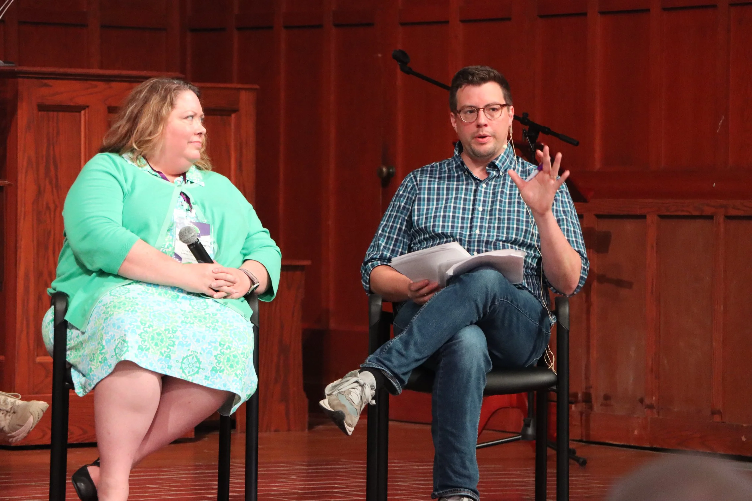 Two people sitting on a stage, engaging in a discussion. The woman on the left holds a microphone, wearing a mint green blazer and skirt, and the man on the right, wearing glasses and a checkered shirt, is speaking and holding papers.