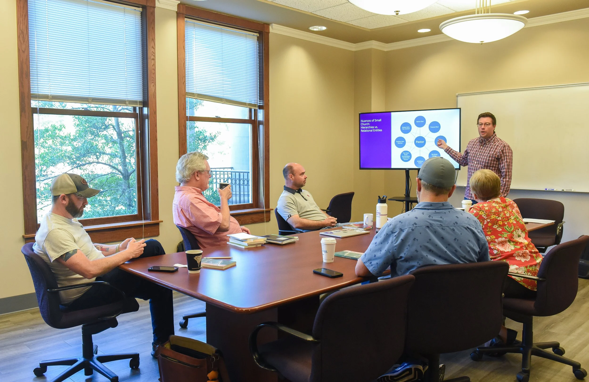 A man in a checkered shirt points to a presentation on a screen in a conference room. Five people are seated around a large wooden table, listening and taking notes. The room has large windows with blinds, and trees are visible outside.