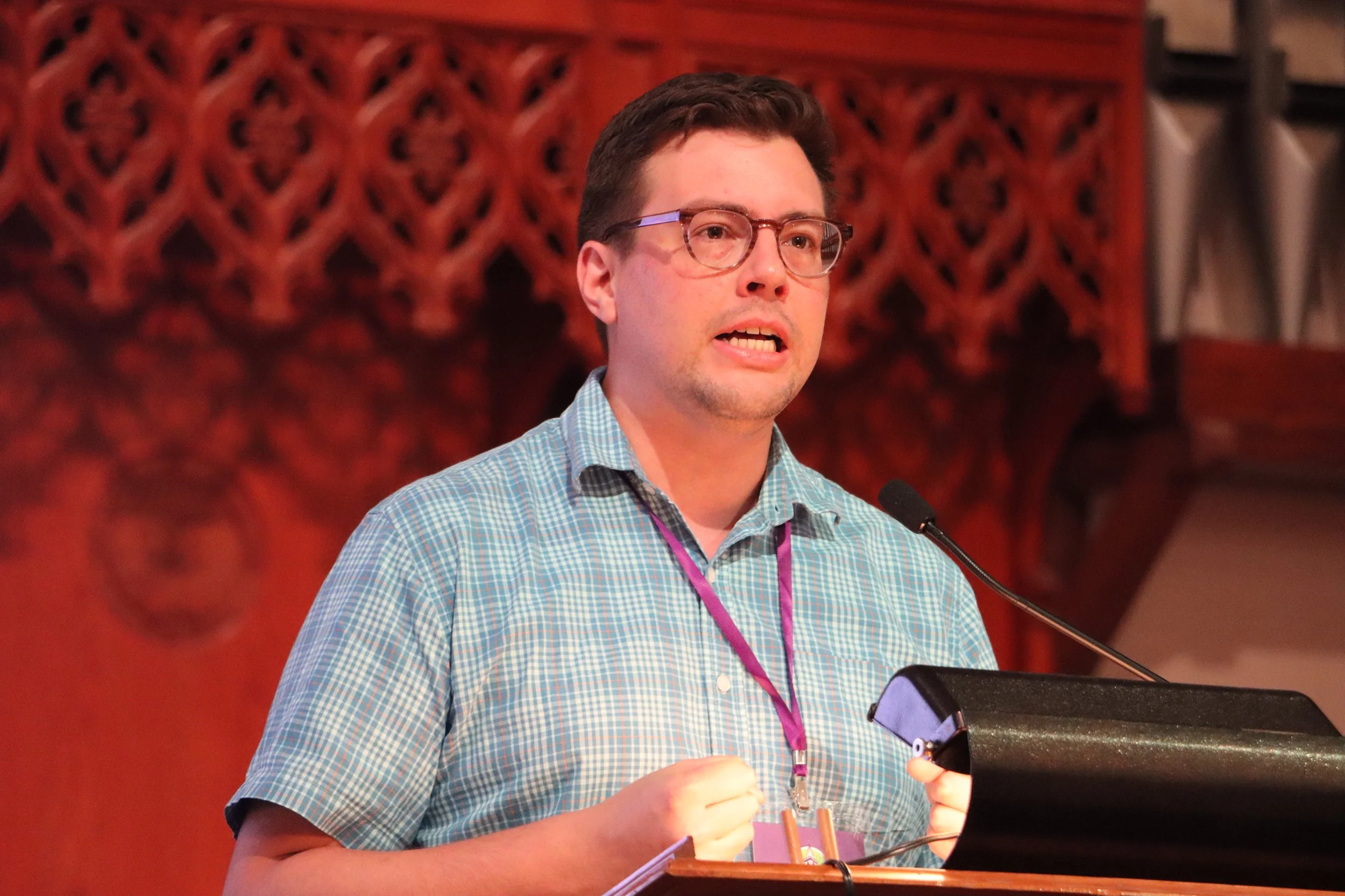 A man with glasses and short dark hair wearing a blue and green checkered shirt, speaking at a podium with a microphone, against a background of wooden latticework.