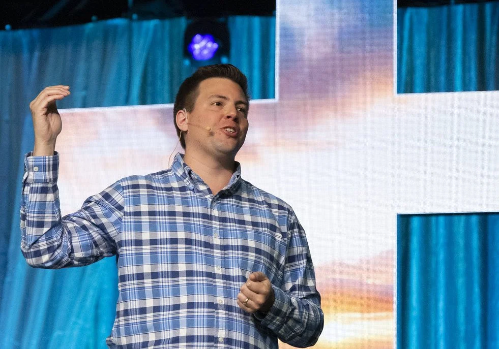 A man giving a presentation on stage with a large screen behind him displaying an abstract or scenic background.