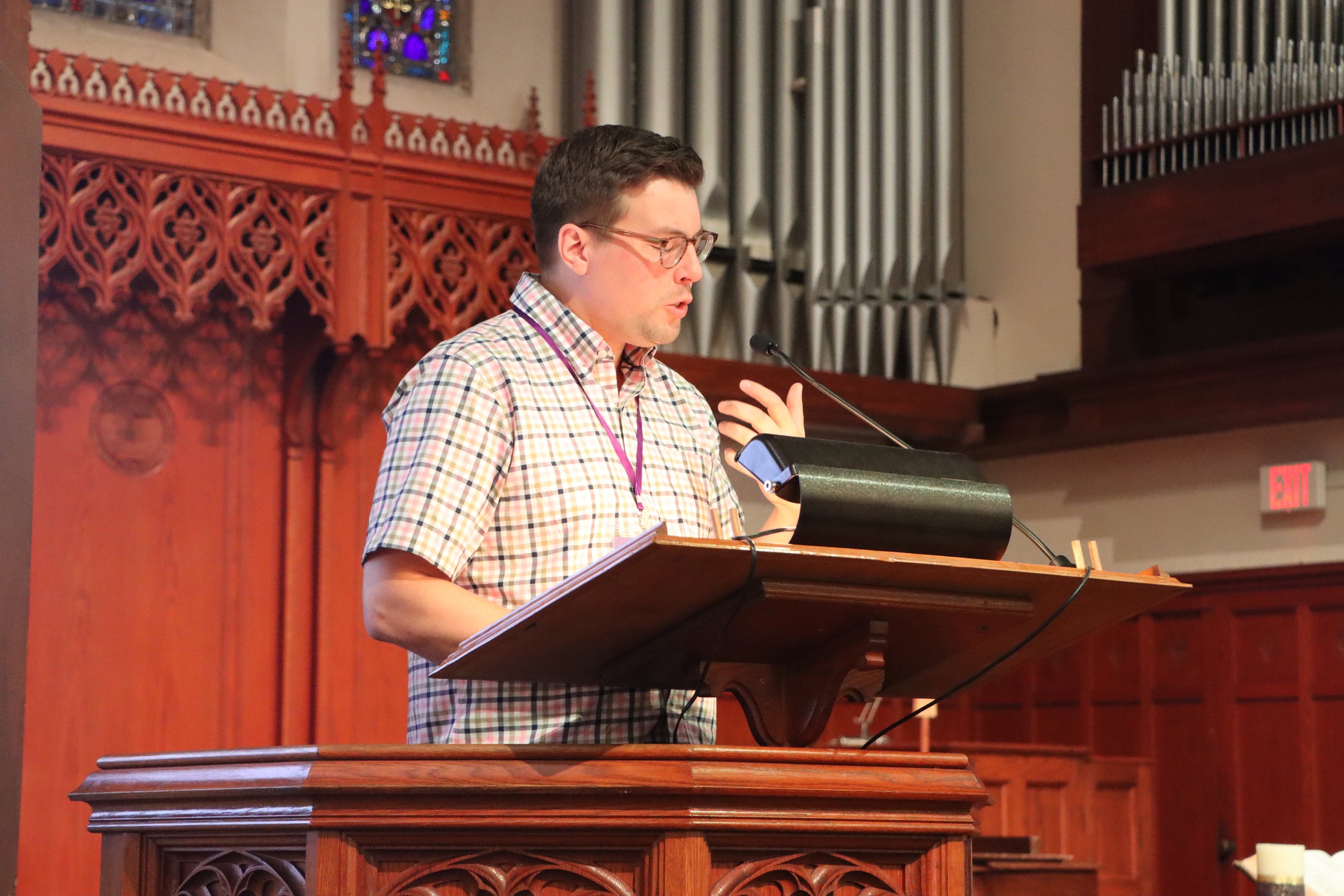 A man in a checkered shirt and glasses speaking at a wooden podium with a microphone in a church or chapel, with stained glass windows and organ pipes in the background.