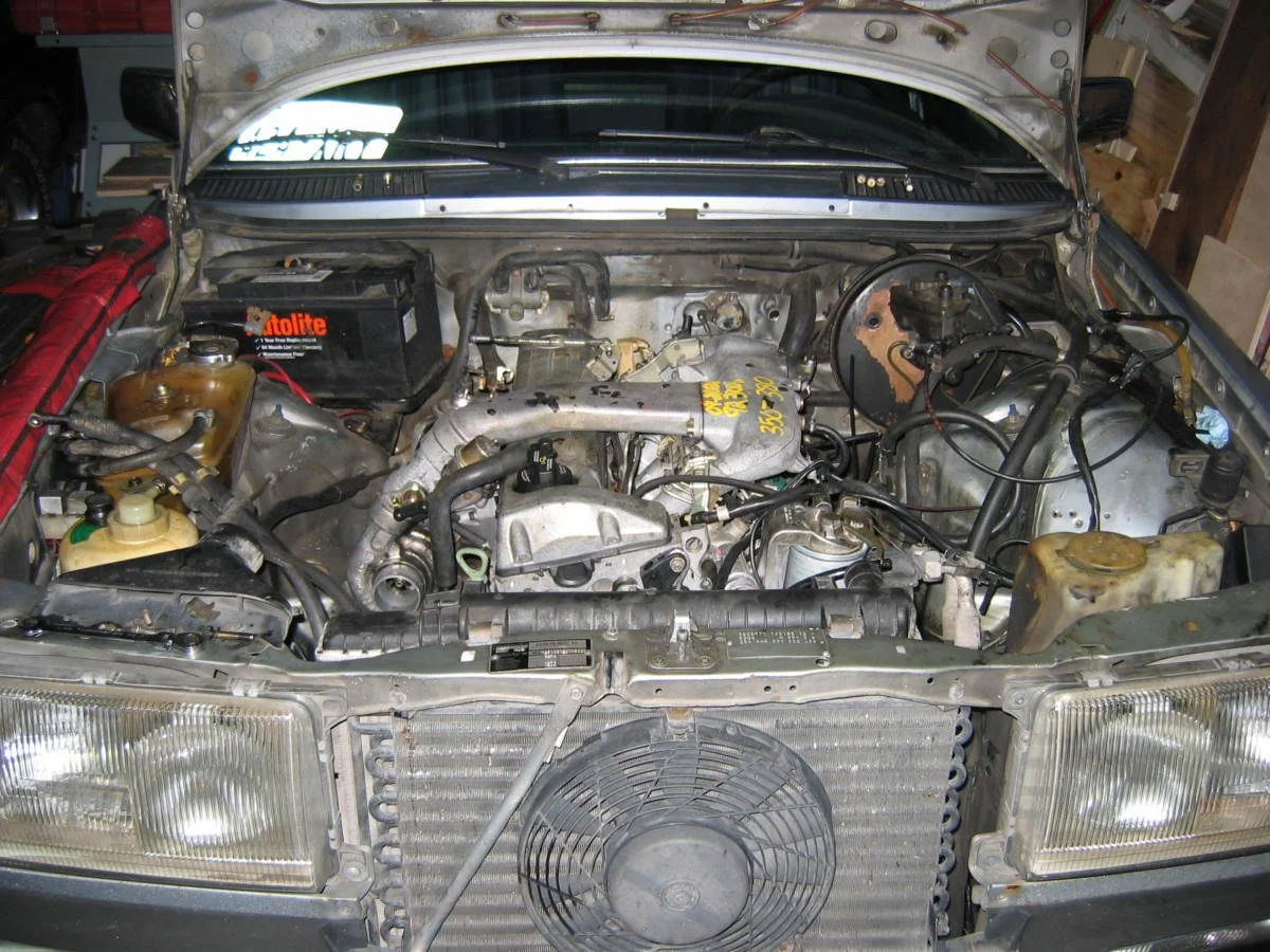Car engine bay with various mechanical parts, radiator at front, battery on the left, with the hood open.