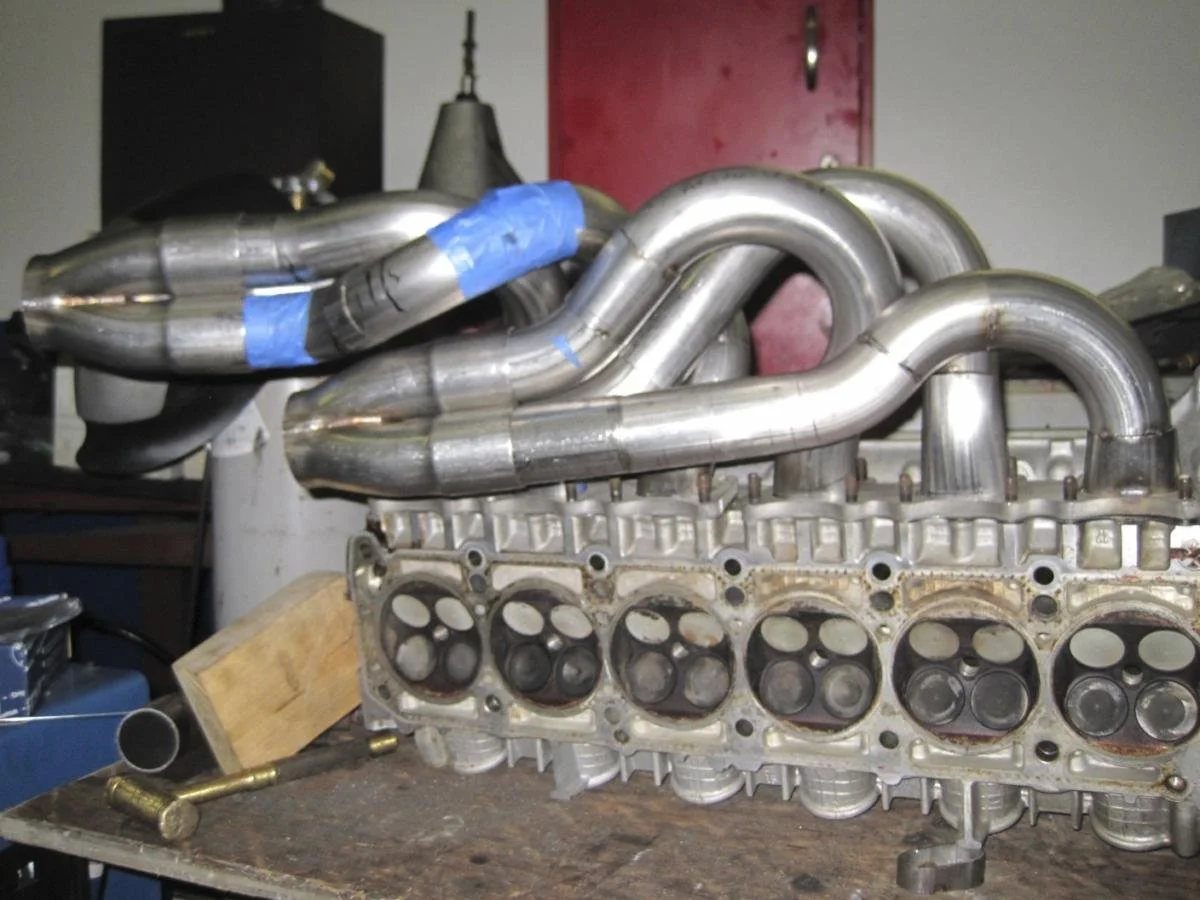 Car engine head with custom exhaust pipes mounted on top, placed on a workbench in a workshop.