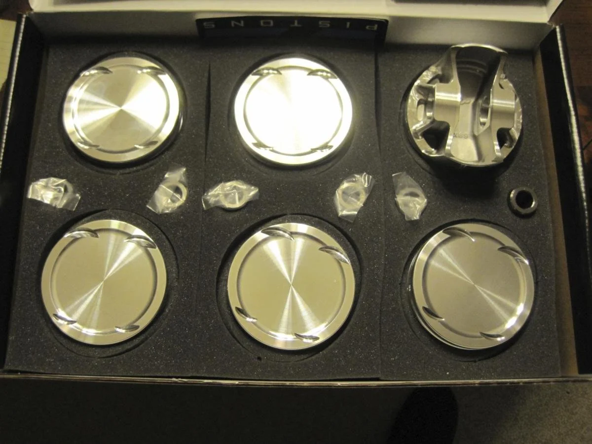 Set of six polished metal pistons in a foam-lined box, some with protective plastic covers.
