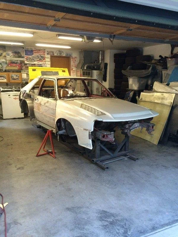 A car body is mounted on a stand inside a garage or workshop, with no wheels or engine installed, possibly under restoration or assembly.