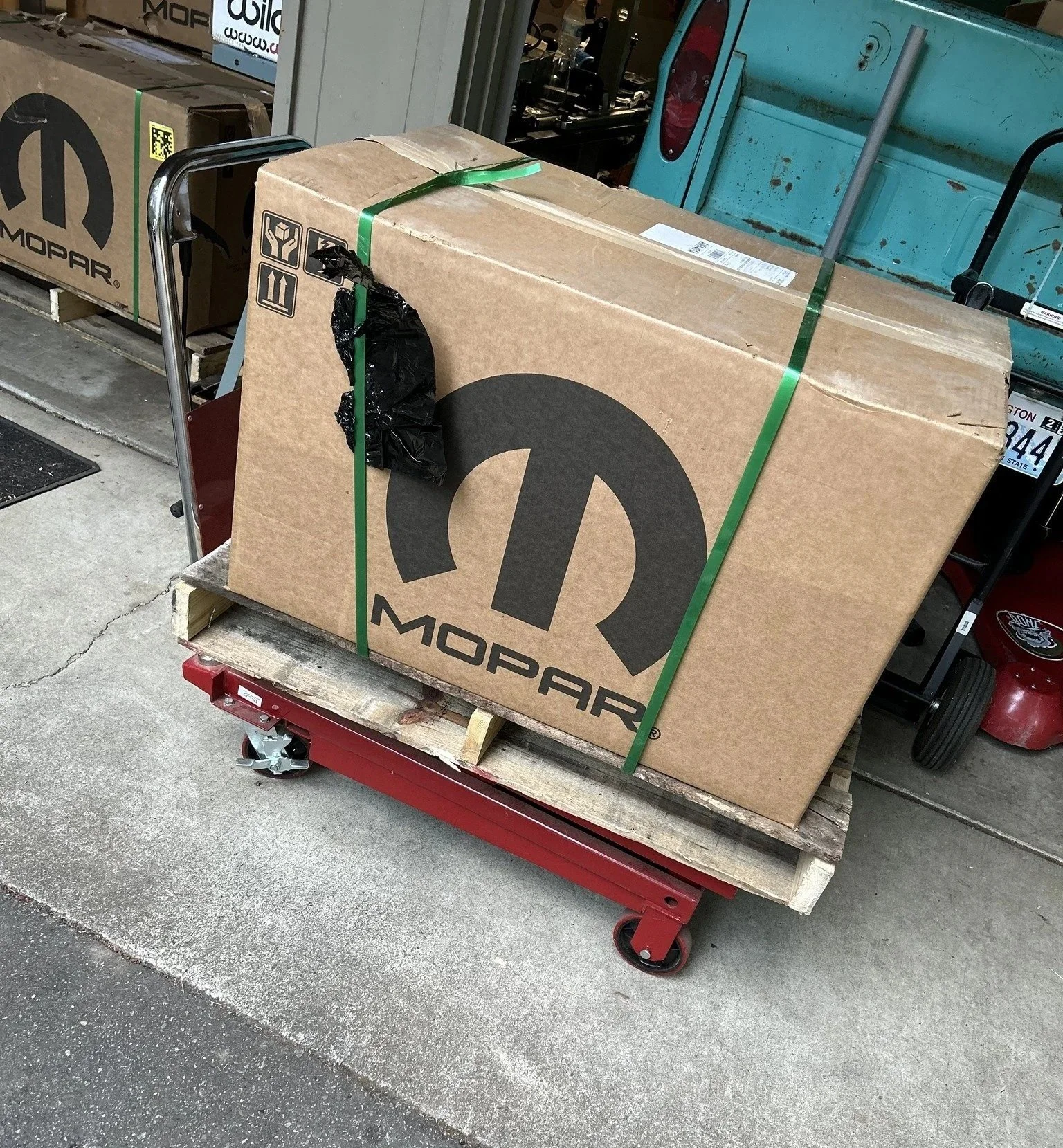 A large cardboard box with the Mopar logo on it, secured with green straps on a red flatbed cart outside a store.