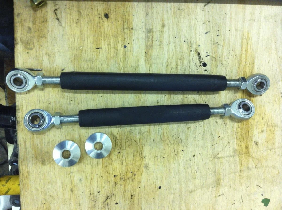 Two motorcycle shock absorbers with metal eyelets and rubber bushings, laid horizontally on a wooden workbench, with two metal washers below them.