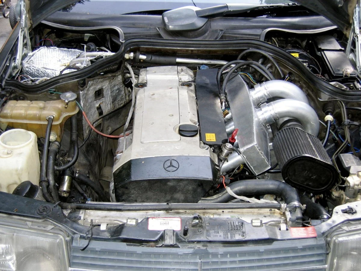 Under the hood of a car, showing the engine with metallic pipes, hoses, and a prominent black air filter, with various engine components and fluid reservoirs visible.