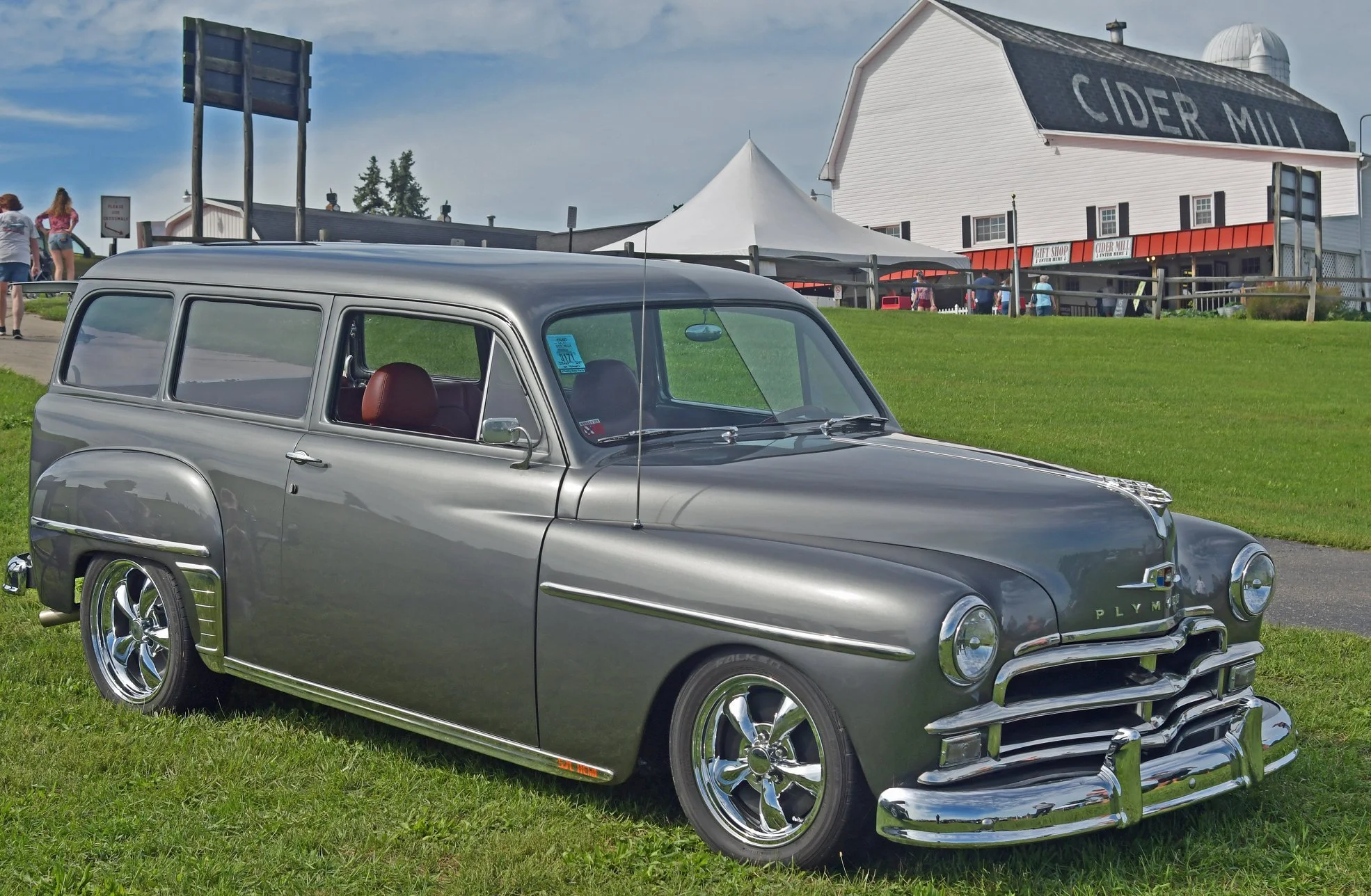 A vintage gray station wagon parked on grass in front of a cider mill. There are people and structures in the background, including a white tent, a building with a sign that says 'CIDER MILL', and other small buildings.