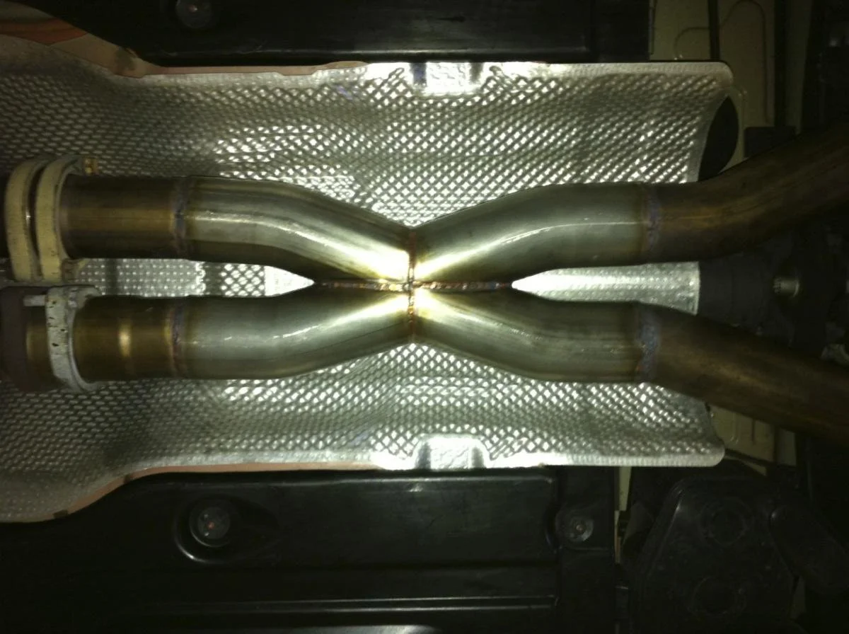 Two metal pipes joined together with welds, placed on a textured metal surface, possibly part of an exhaust system or similar mechanical component.