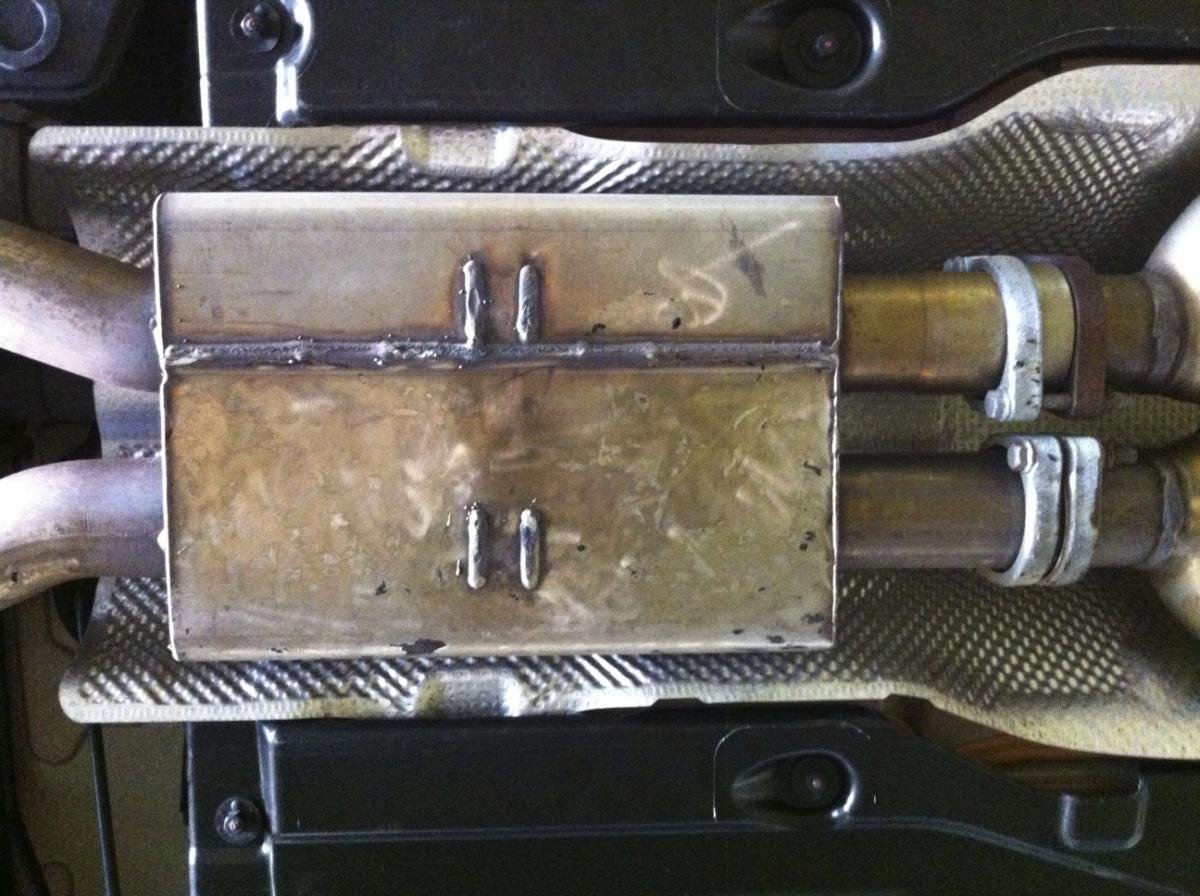 Two metal pipes connected to a metal box with welds, mounted on a textured surface.