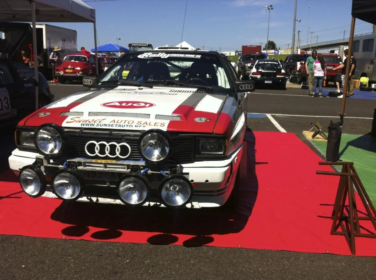 A vintage Audi rally car on display at a car show, featuring sponsor logos and multiple large rally lights mounted on the front, parked on a red carpet with tents and other cars in the background.