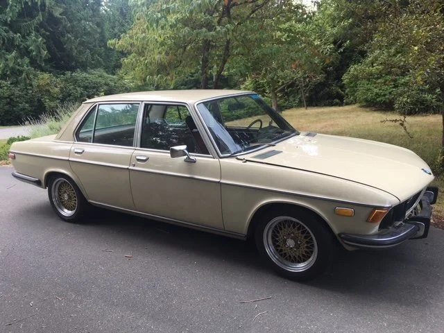 A vintage beige four-door sedan parked on a shaded street with trees in the background.