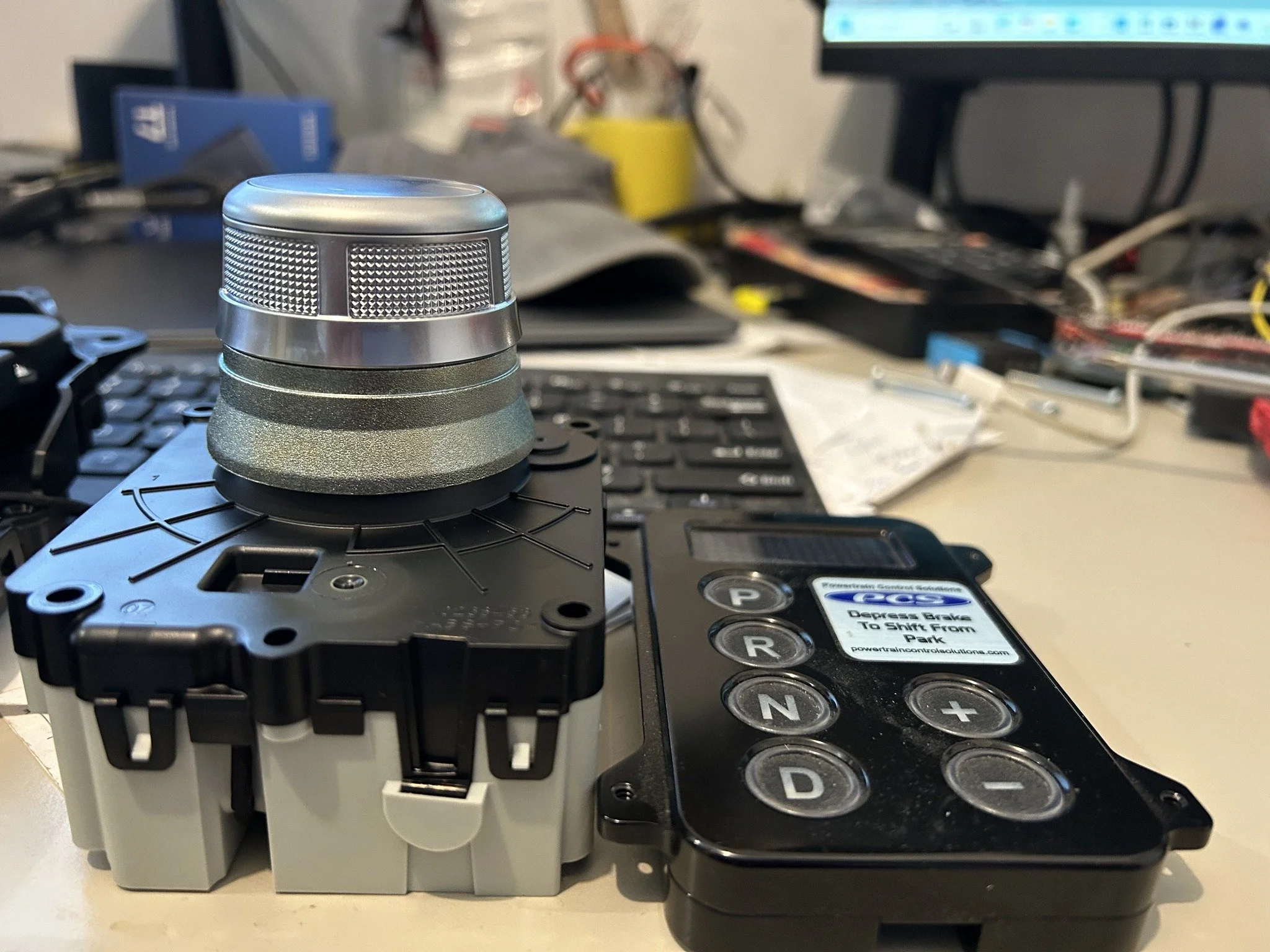 A metallic rotary knob mounted on a black mechanical device, with a remote control labeled 'Depress Brake To Shift From Park' next to it, on a cluttered desk with computers and wires in the background.