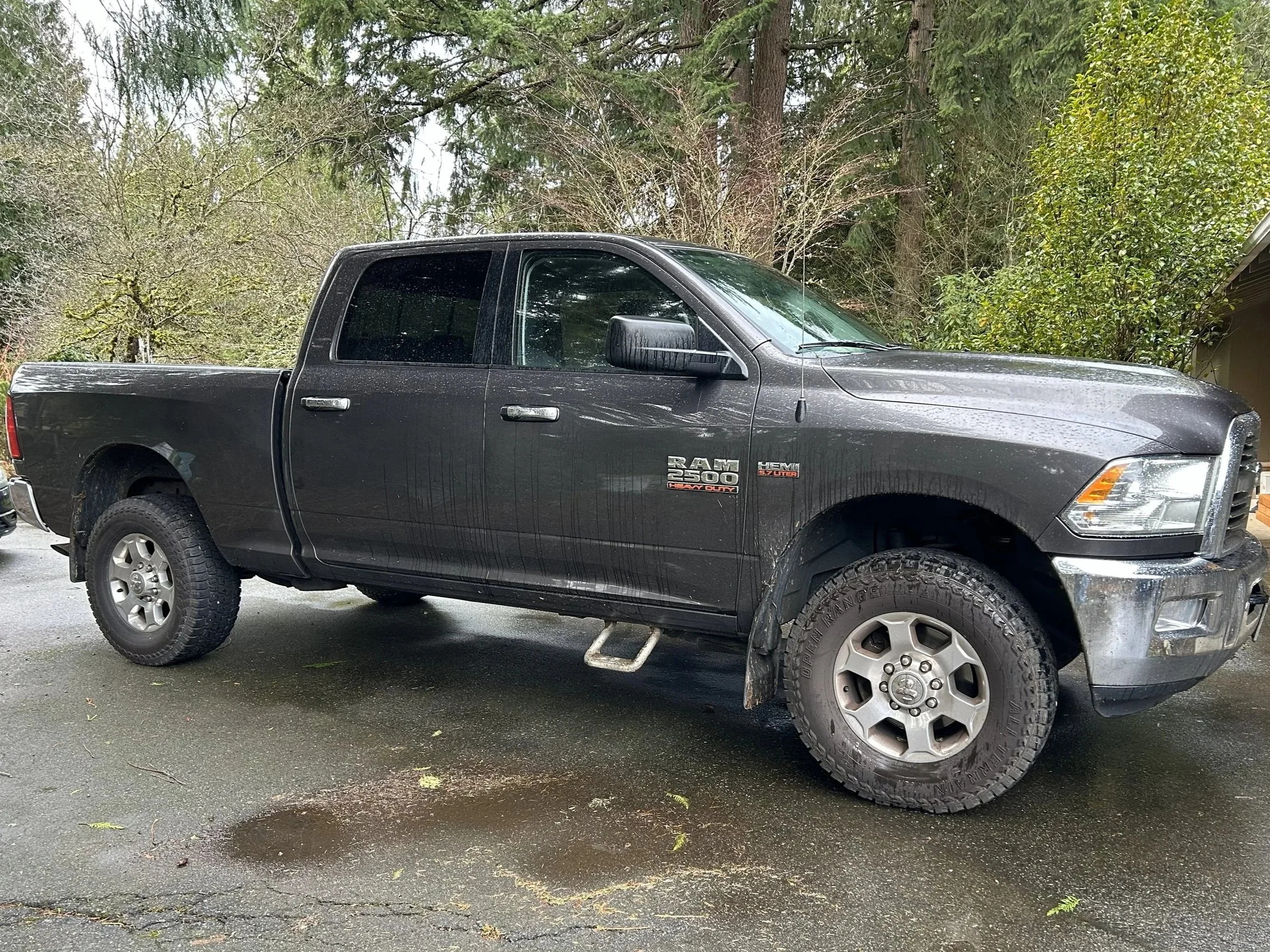 Black Ram 2500 pickup truck parked on wet pavement with mud splatters, surrounded by trees and forested area.