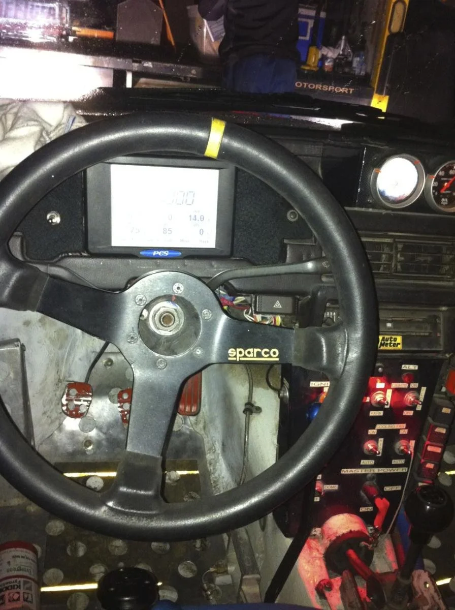 Inside a race car cockpit, showing a black Sparco steering wheel, a digital display screen, and various switches and gauges on the dashboard.