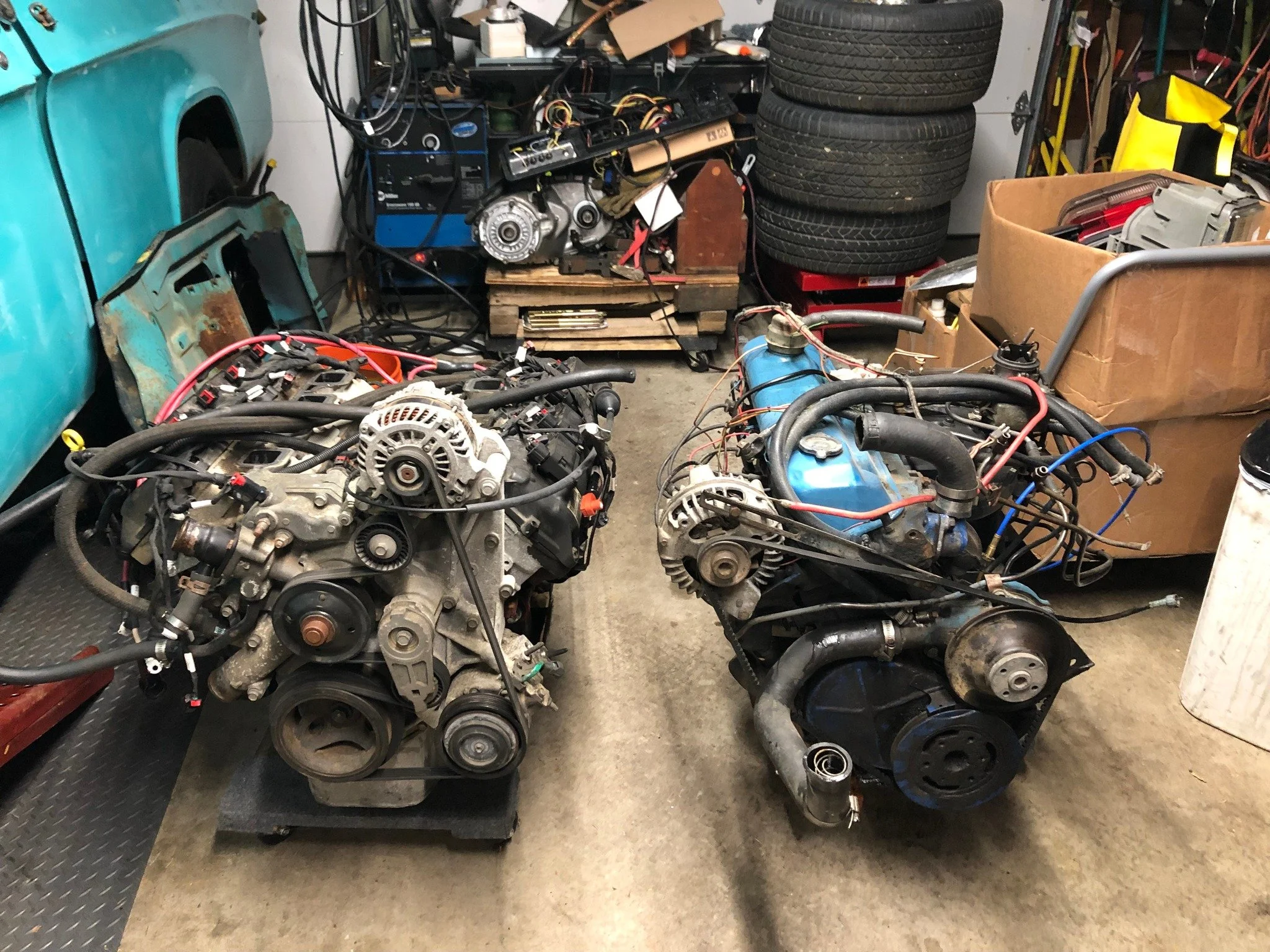 Two car engines on stands in a cluttered garage. The background shows tires, boxes, and tools.