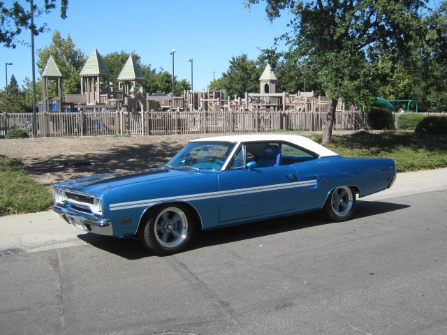 A vintage blue muscle car with a white roof parked on a street, with a playground and trees in the background.