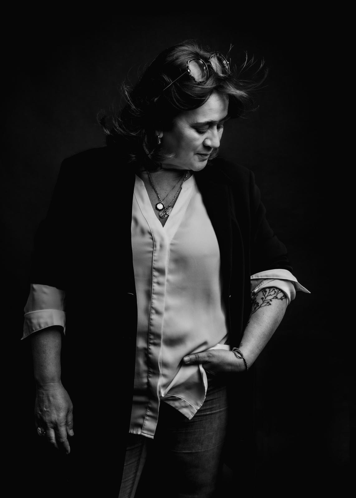Portrait en noir et blanc d'une femme avec des lunettes sur la tête, portant une chemise en satin et une veste, regardant vers le bas, avec un tatouage au bras et des bijoux.