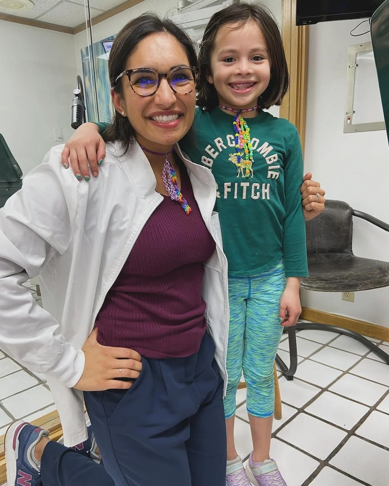 A woman and a young girl smiling, posing together indoors. The woman has dark hair, glasses, and is wearing a white lab coat over a purple shirt, and colorful jewelry. The girl has shoulder-length dark hair, braces, and is wearing a green Abercrombie & Fitch shirt, colorful striped leggings, and a colorful beaded necklace.