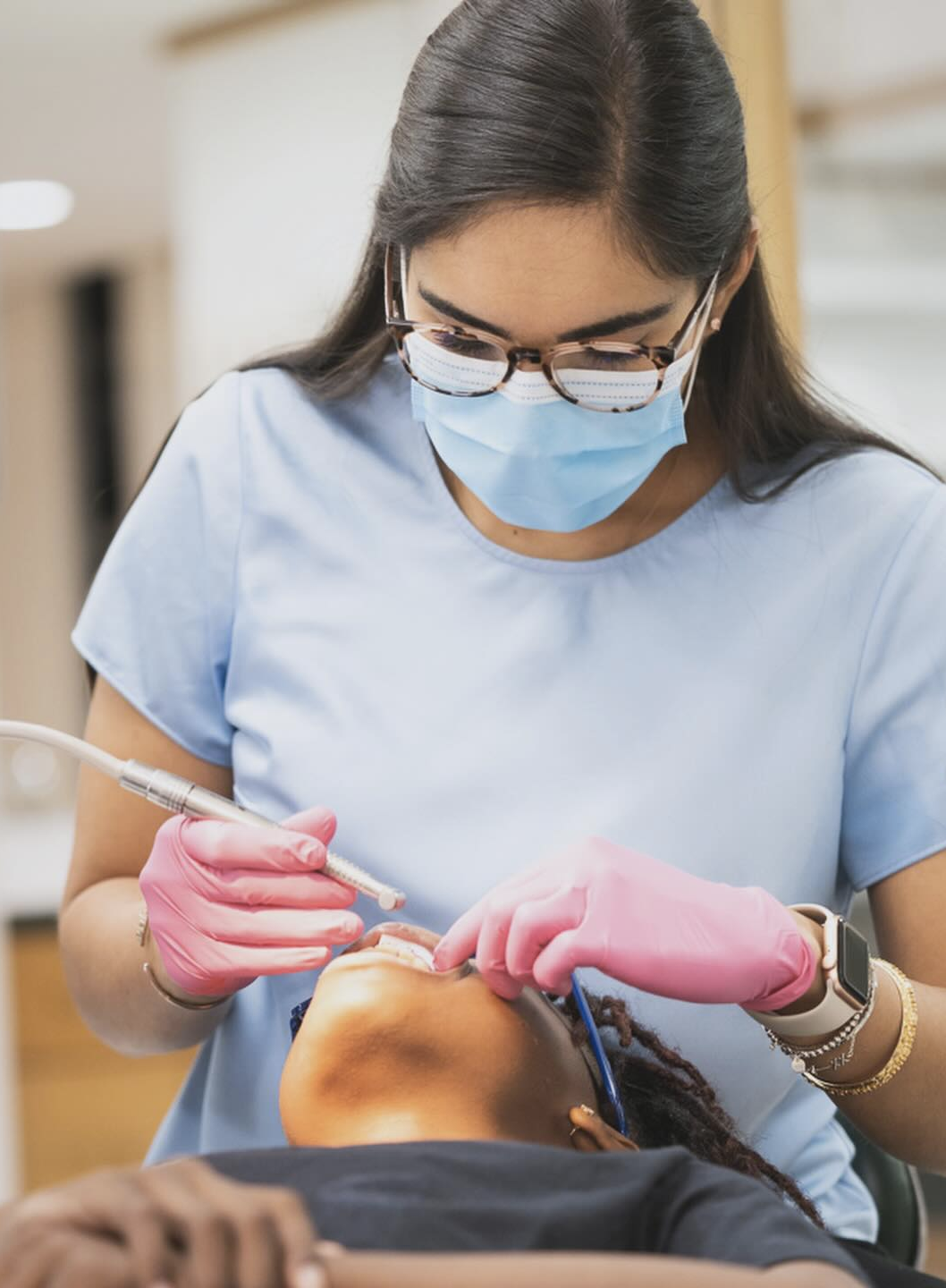 A dental professional wearing glasses, a mask, pink gloves, and a light blue uniform performs a dental procedure on a patient in a dental clinic.