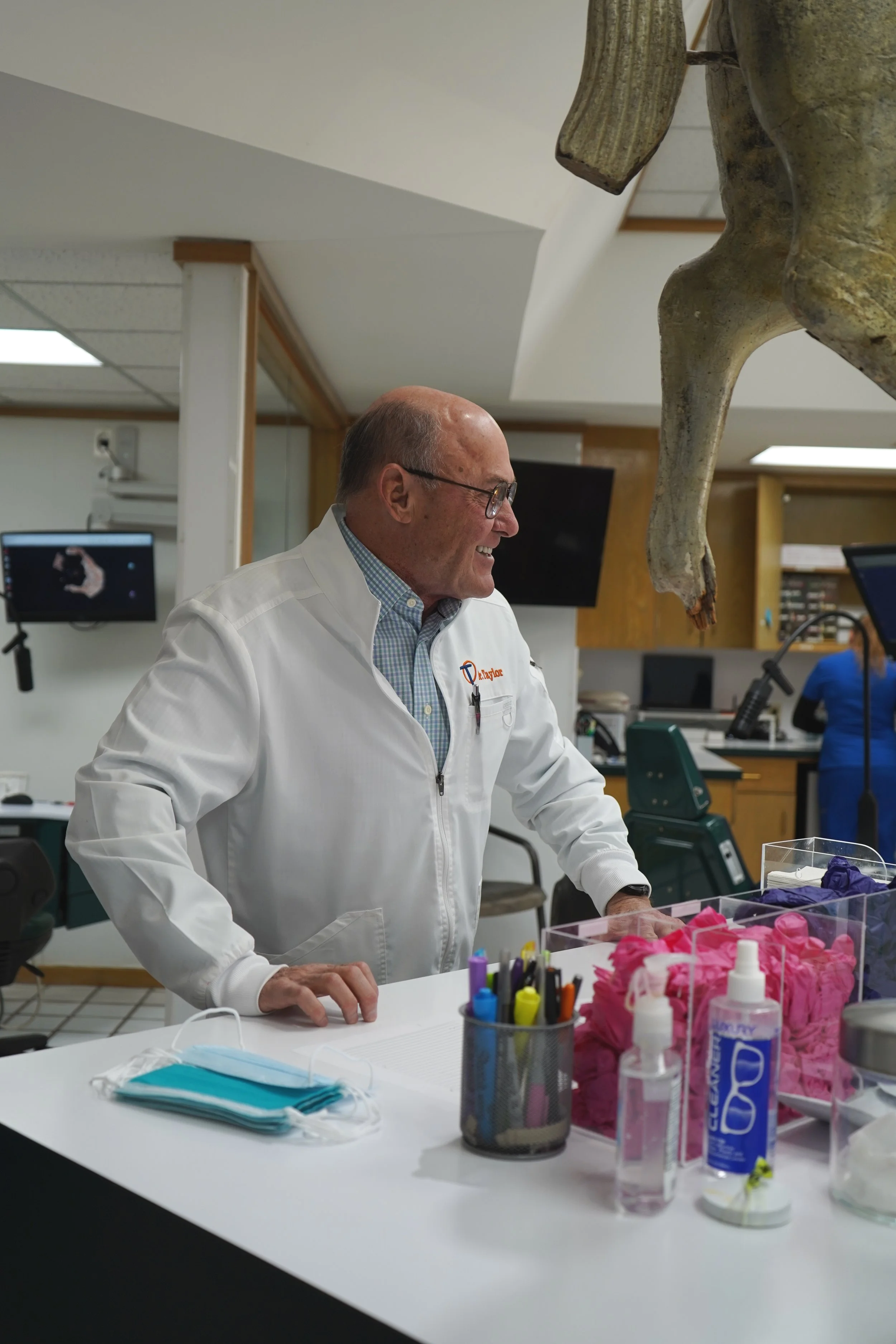 A man in a white lab coat standing at a laboratory counter, smiling, with medical masks, colorful pens, and sanitizer on the table. A horse skull is hanging above him.