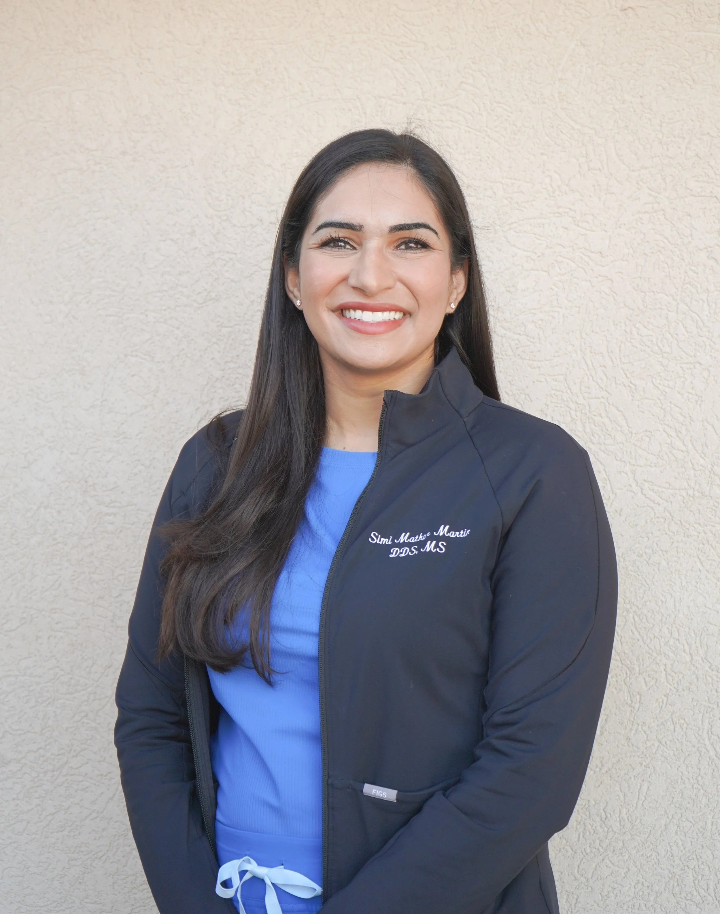A smiling woman with long dark hair wearing a blue scrub top and a black jacket with her name and credentials embroidered on it, standing against a beige wall.