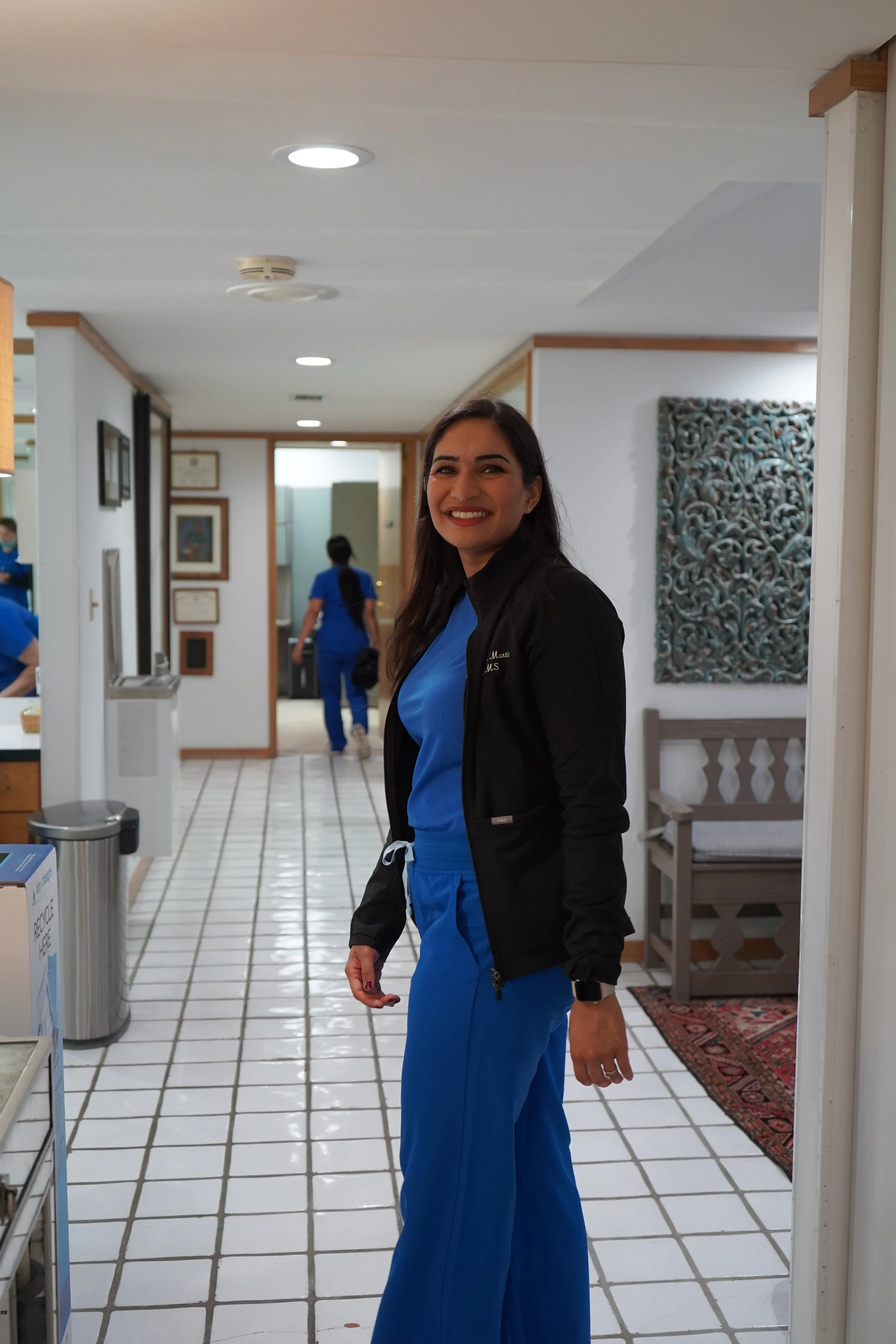 Dr. Mathur wearing blue medical scrubs and a black jacket standing in a corridor of a medical facility, with two other women in blue scrubs walking away in the background.