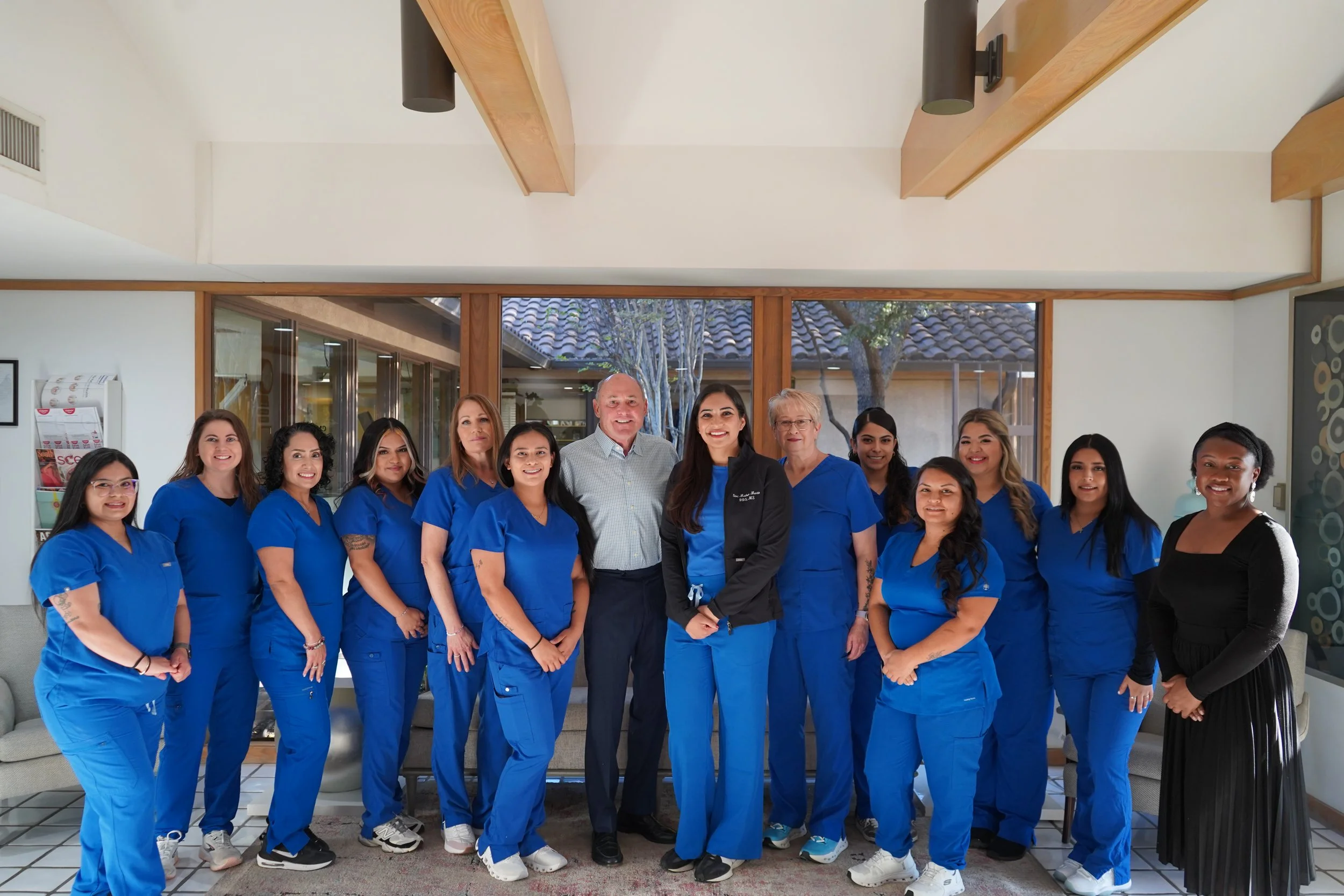 A group of healthcare professionals, mostly women, wearing blue scrubs, standing together with a man and a woman in the middle, inside a well-lit room with large windows and wooden trim.
