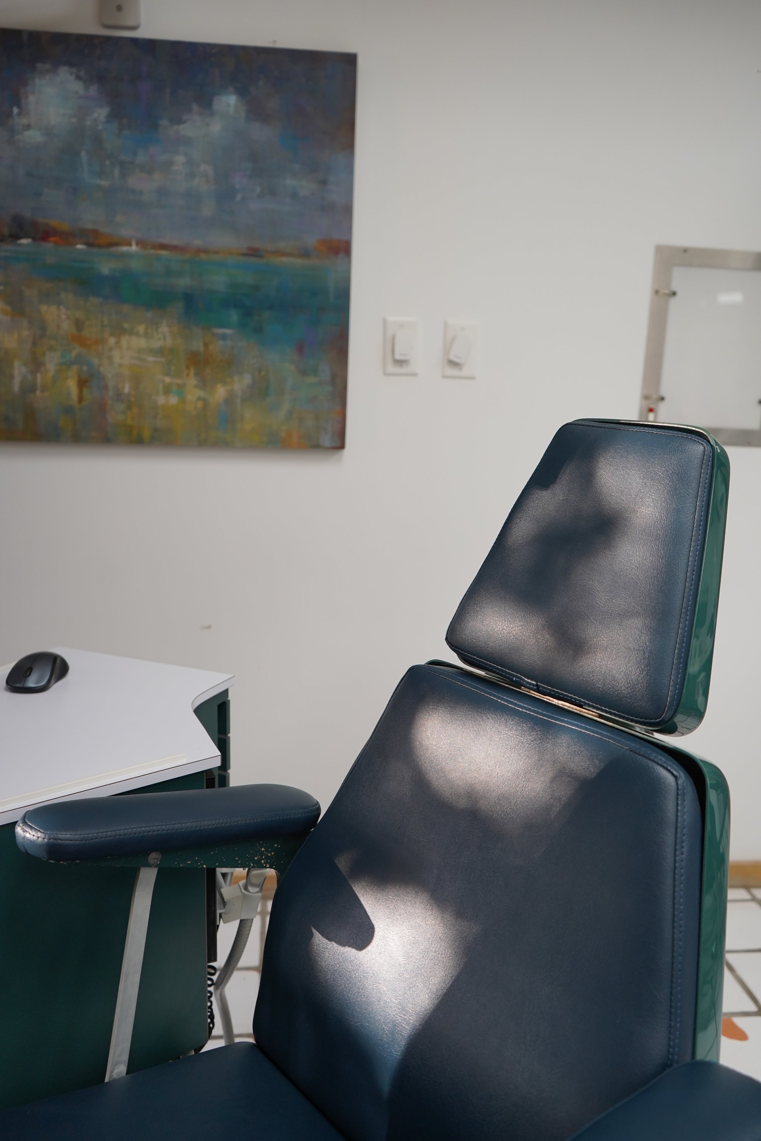 An empty medical examination room with a medical chair, a white desk with a computer mouse, a large abstract painting on the wall, and a whiteboard frame.