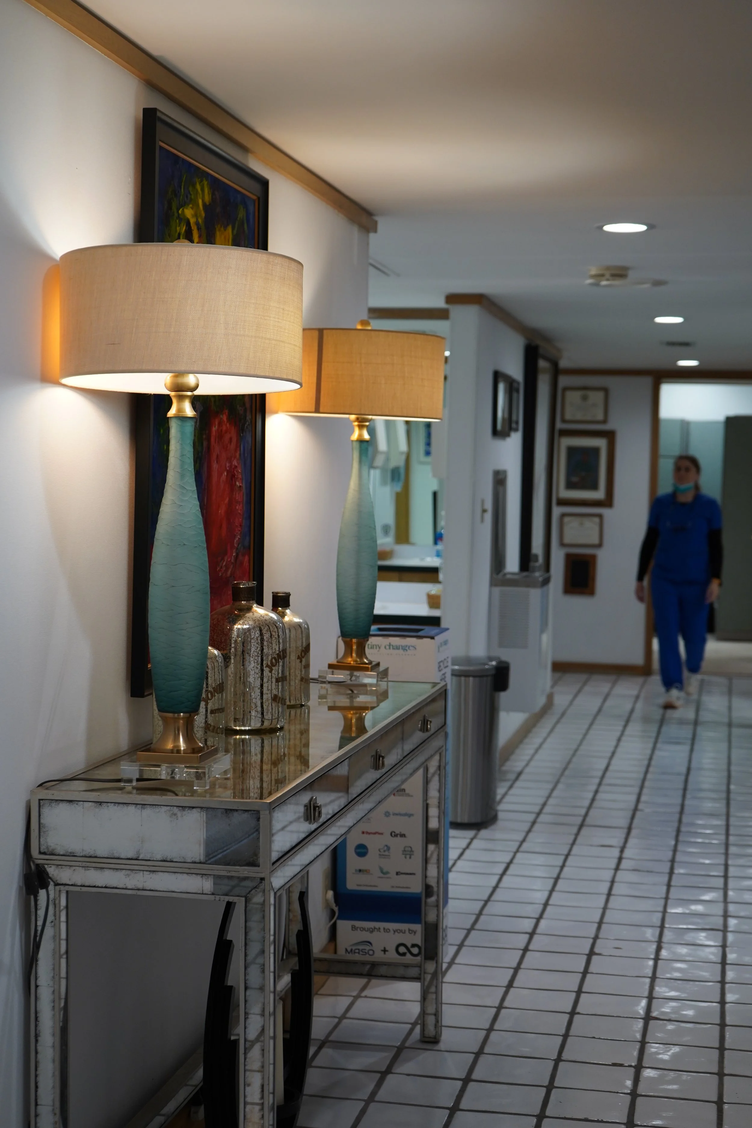 A hallway with two blue and gold table lamps on a white marble console table, with framed artwork on the wall behind them. In the background, a woman in blue scrubs is walking down the hallway.