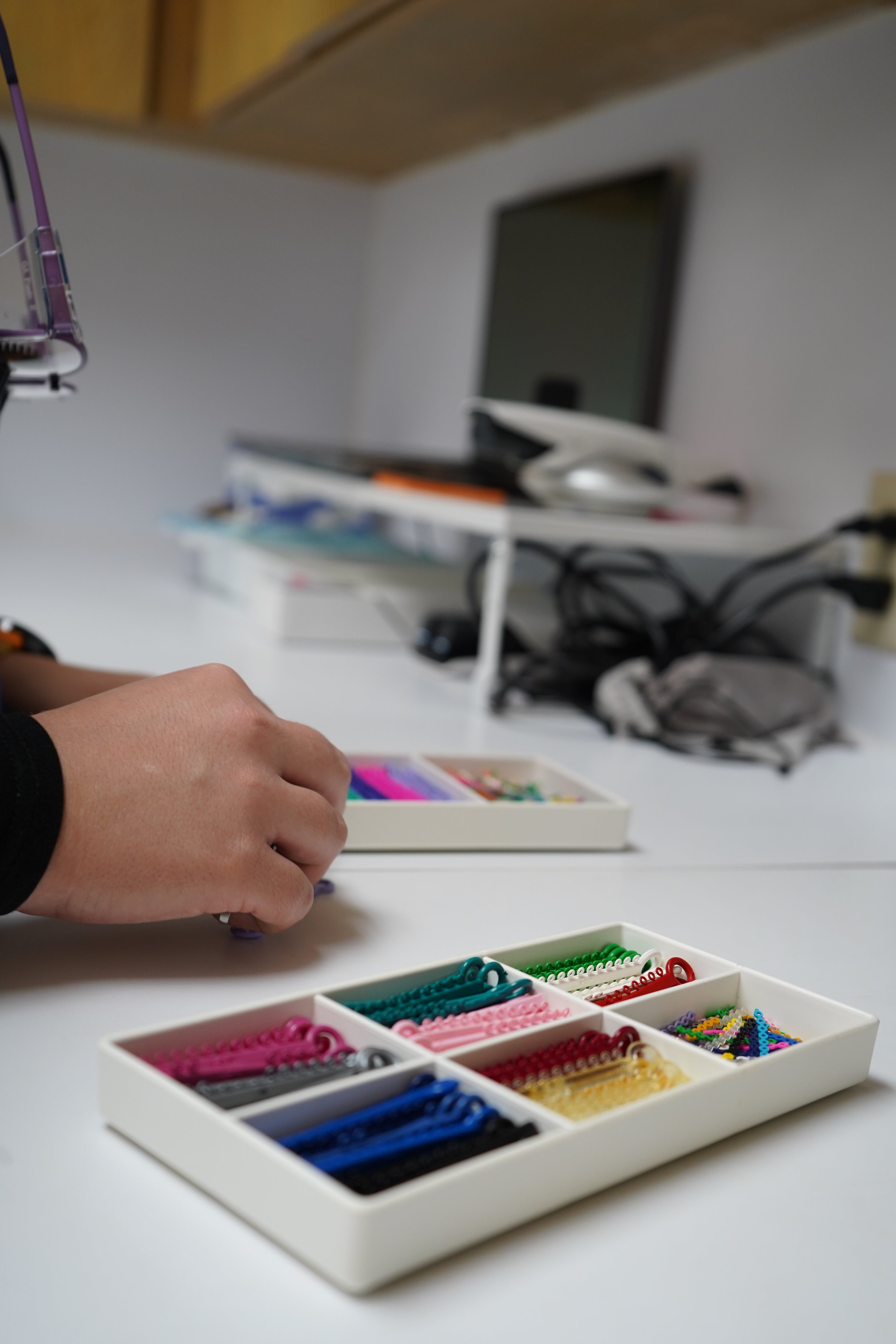 A hand reaching for colorful hair accessories organized in white drawers on a white desk.