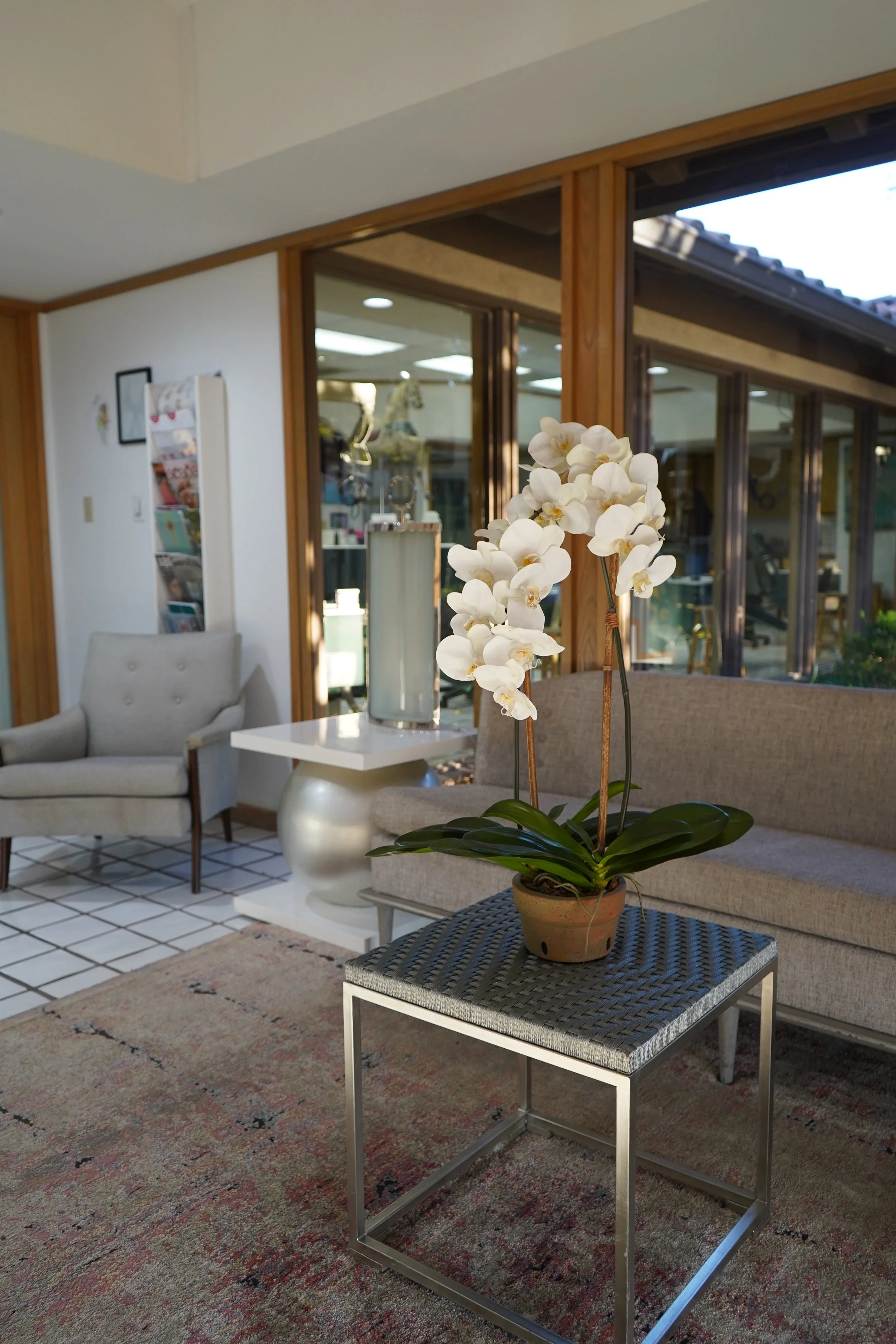 A living room with a potted white orchid on a small table, beige and gray furniture, and large windows with wooden frames.